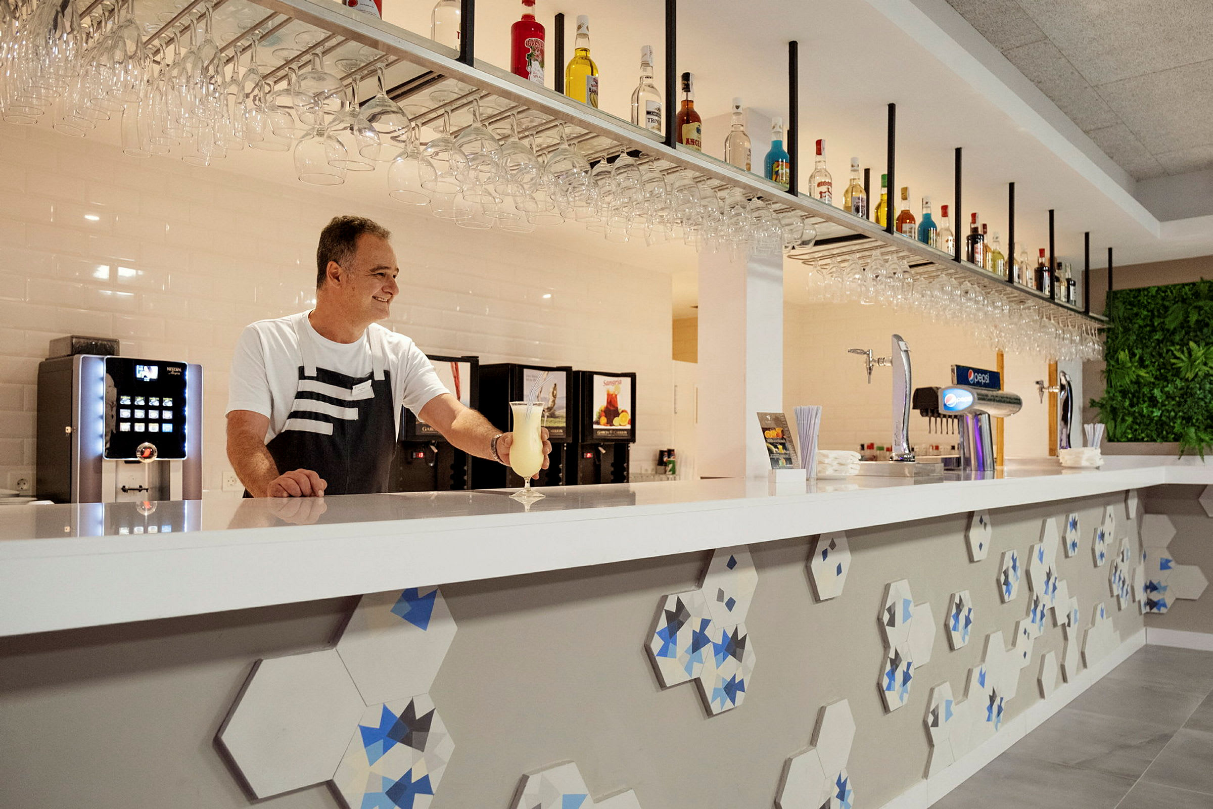 En bartender ved en moderne bardisk med flasker og glass hengende ovenfor. (AI-generert tekst)