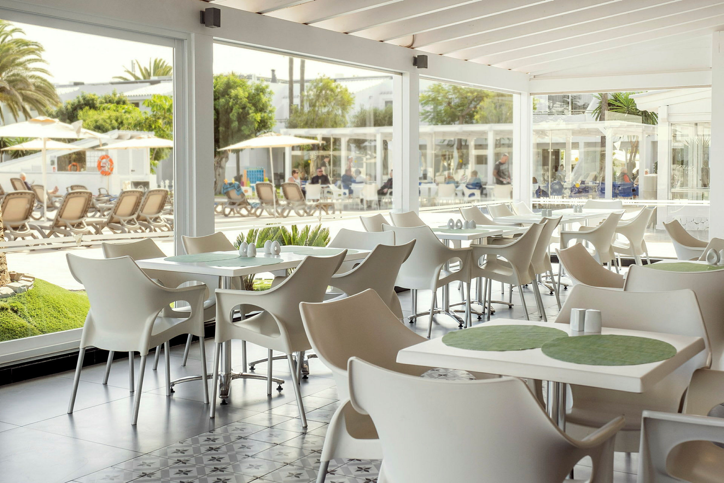 En lys og moderne innredet kafé med hvite stoler og bord, med utsikt mot utendørs bassengområde gjennom store glassvinduer. (AI-generert tekst)