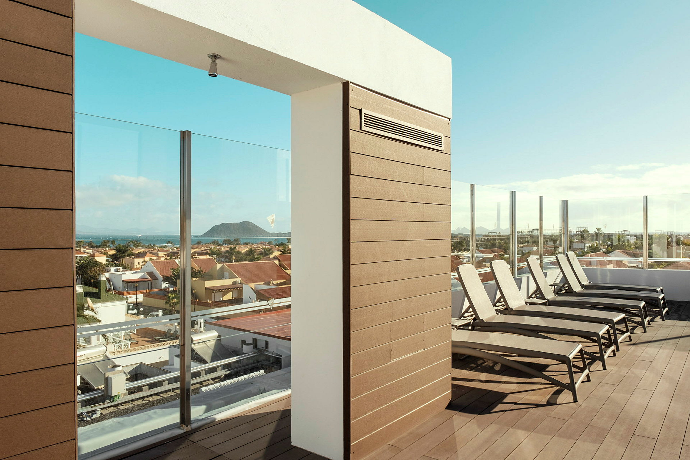 En solterrasse med solsenger og glasrekkverk, med utsikt over et boligområde og en fjern ås. (AI-generert tekst)