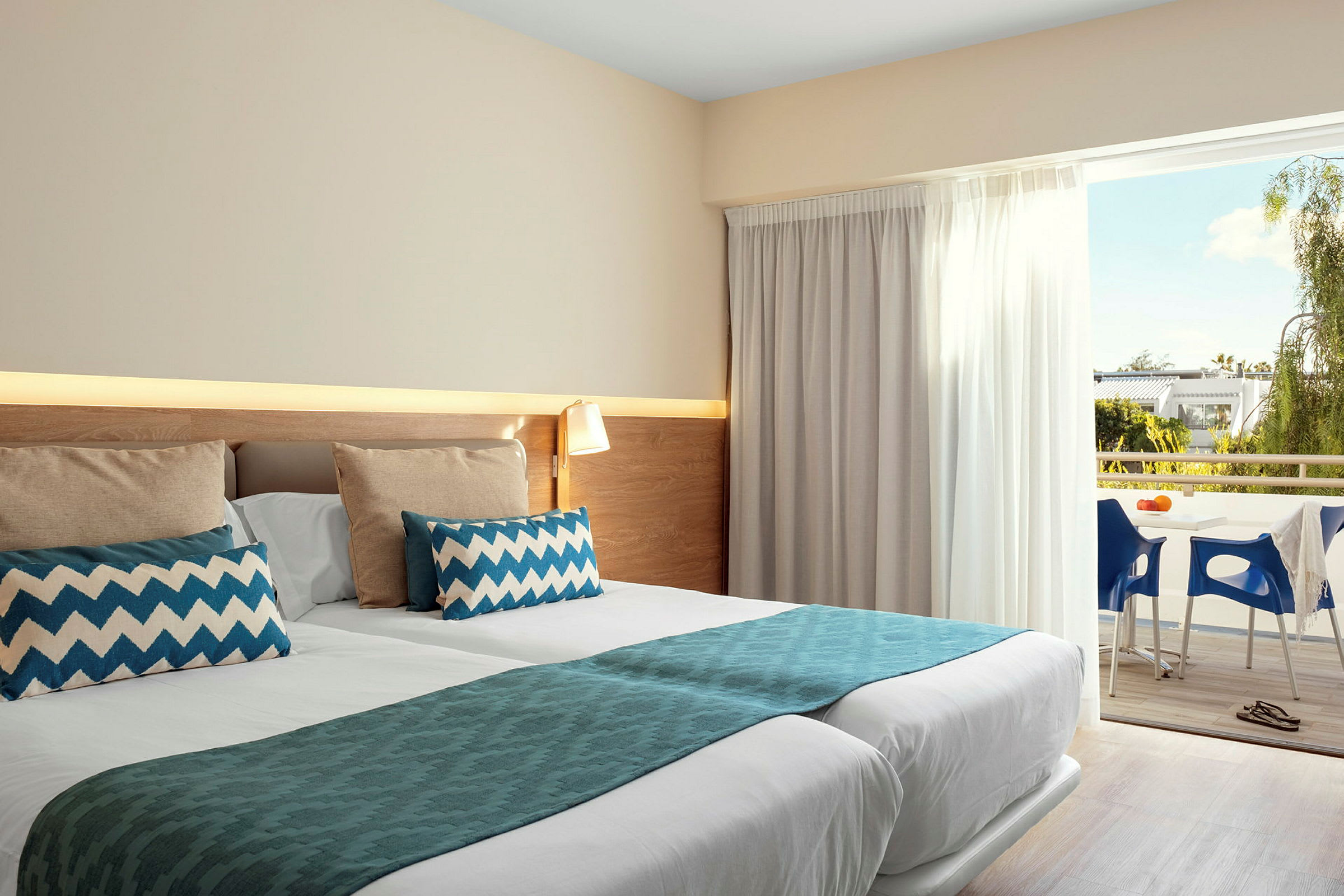Et hotellrom med dobbeltseng som er pent oppredd med blå detaljer og puter. Rommet har også tilgang til en balkong med utsikt over grøntområder og møbler. (AI-generert tekst)