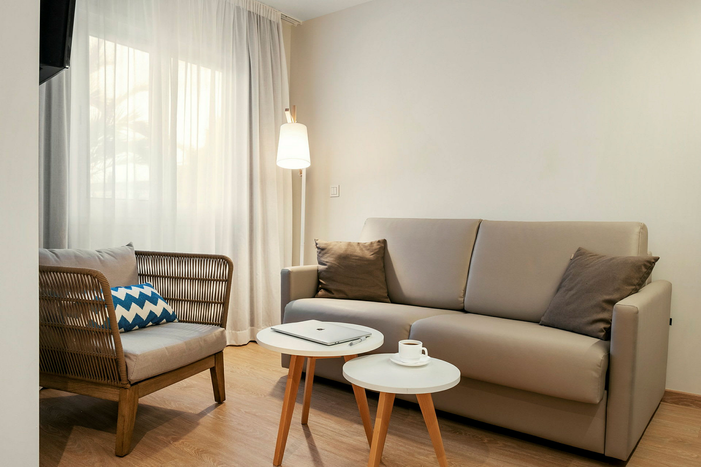 En stue med en beige sofa, en stol med flettet rygg, to små hvite bord og en gulvlampe. Et vindu med lyse gardiner er synlig i bakgrunnen. (AI-generert tekst)