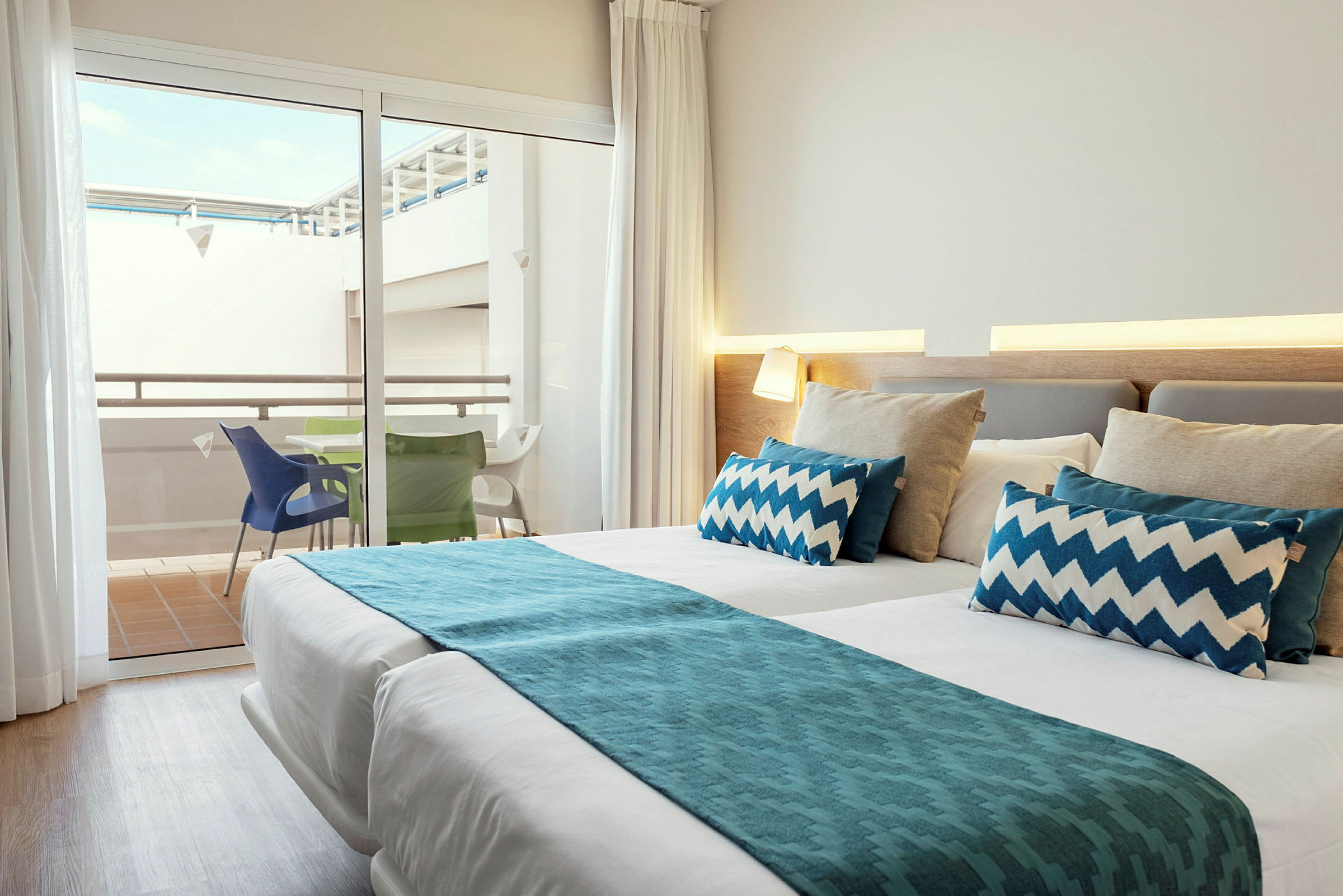 Et lyst og moderne hotellrom med en oppredd dobbeltseng og en uteplass synlig gjennom glassdørene, hvor det står et bord med fargerike stoler. (AI-generert tekst)