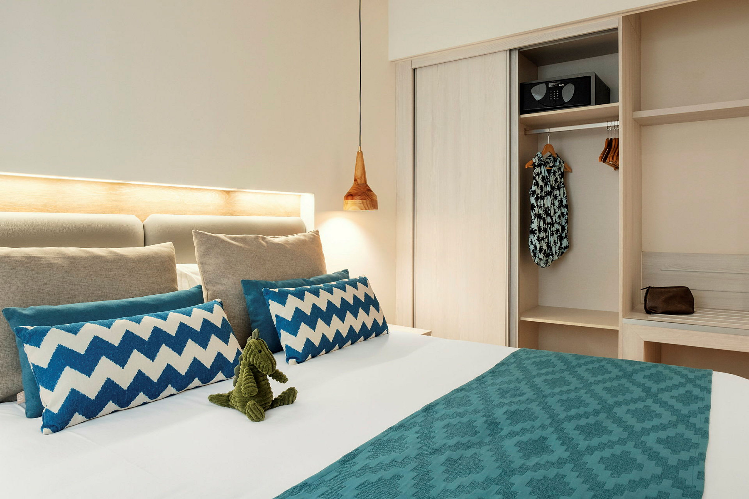 Et hotellrom med en oppredd seng som har dekorative blå puter og en grønn lekefigur. Et skap med klær og en safe er synlig ved siden av. (AI-generert tekst)