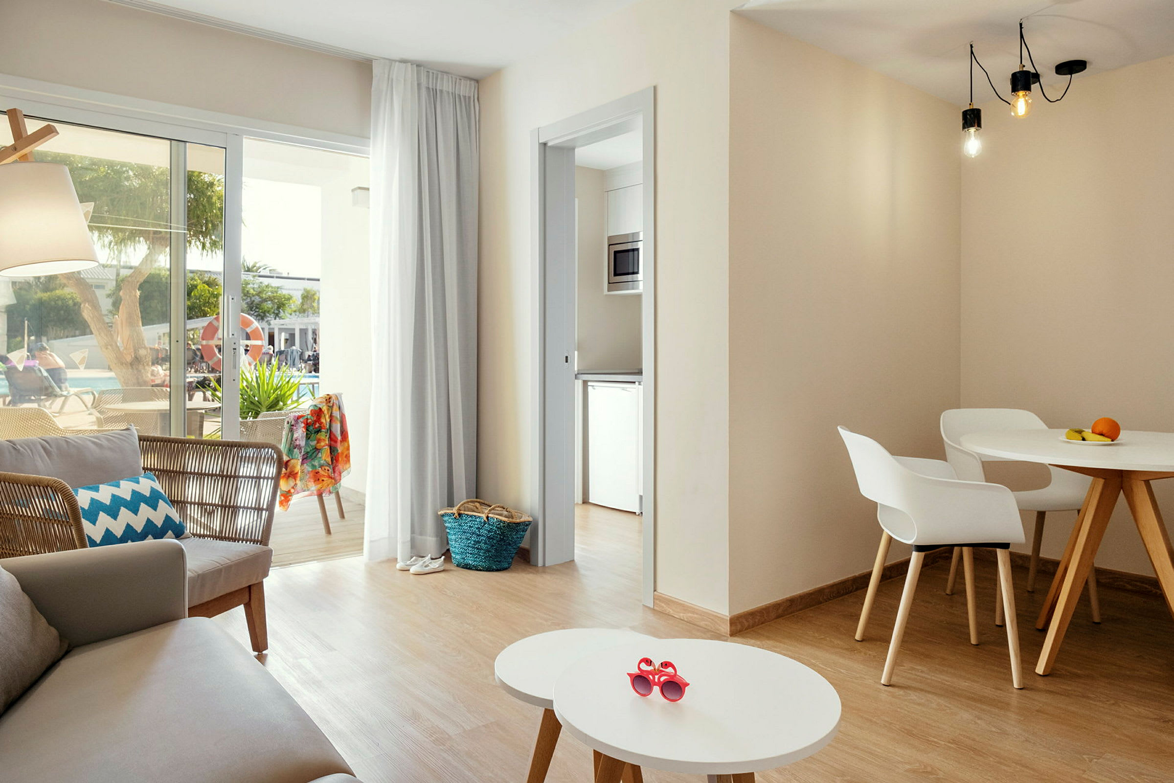 En lys og moderne innredet stue med sittegruppe, spisebord og store vinduer som vender mot en terrasse med utsikt over et basseng. (AI-generert tekst)