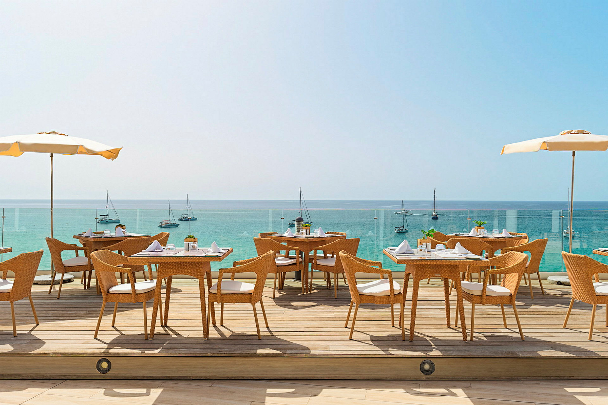 Utendørs restaurant med utsikt over havet. Flere bord og stoler står på en terrasse med parasoller, og seilbåter er synlige i bakgrunnen. (AI-generert tekst)