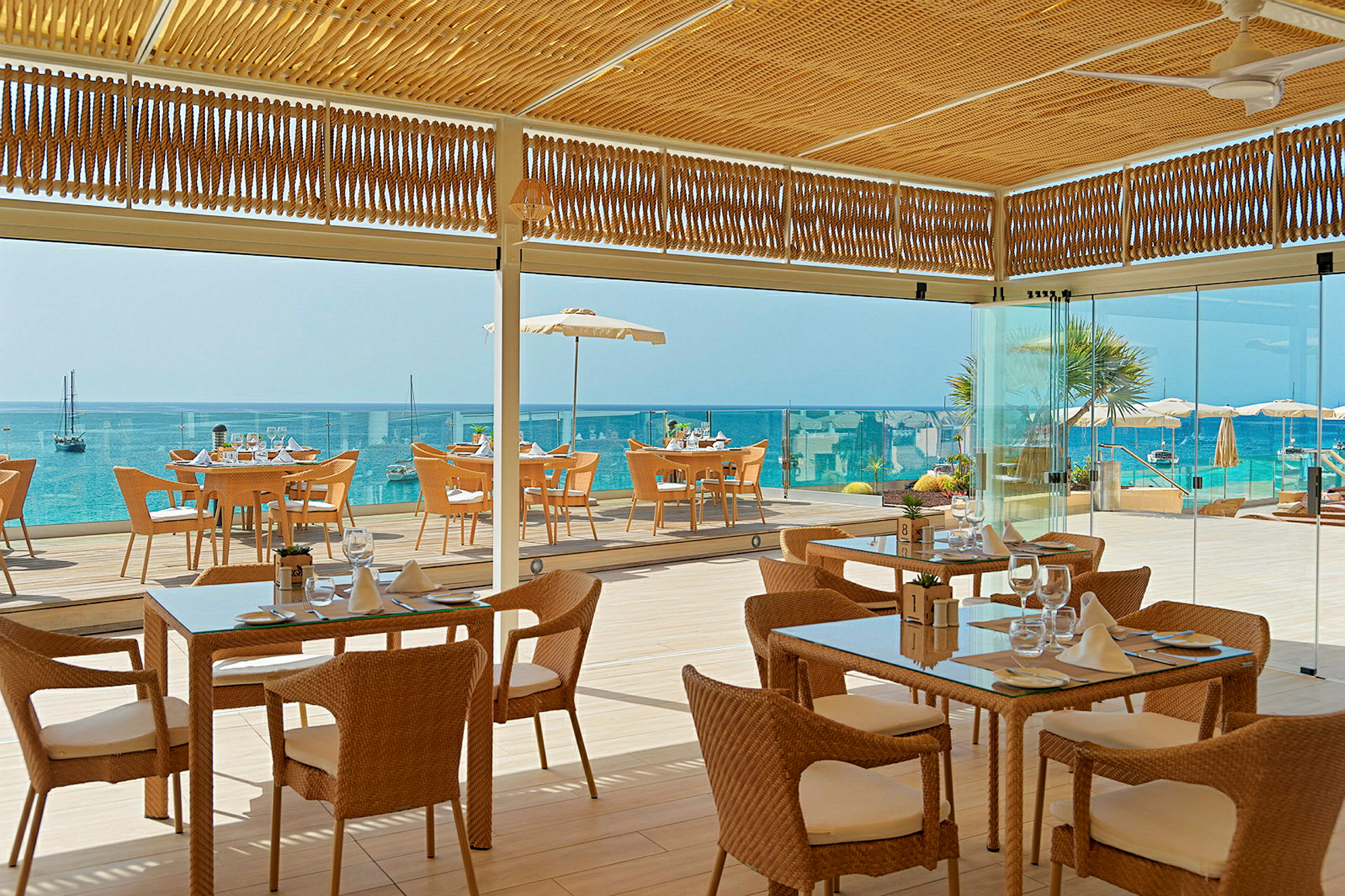 Bilde av en restaurant med flettede stoler og glassbord, beliggende ved havet med utsikt mot en blå vannhorisont. (AI-generert tekst)