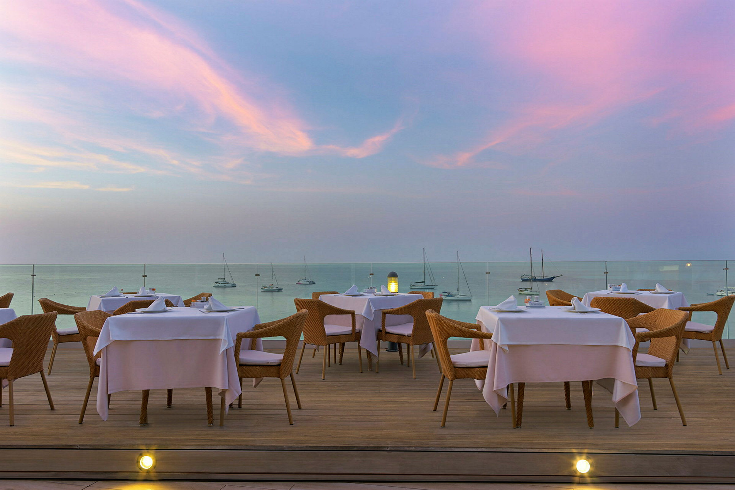 En restaurantterrasse med bord og stoler, med utsikt over havet under en rosa og blå kveldshimmel. (AI-generert tekst)