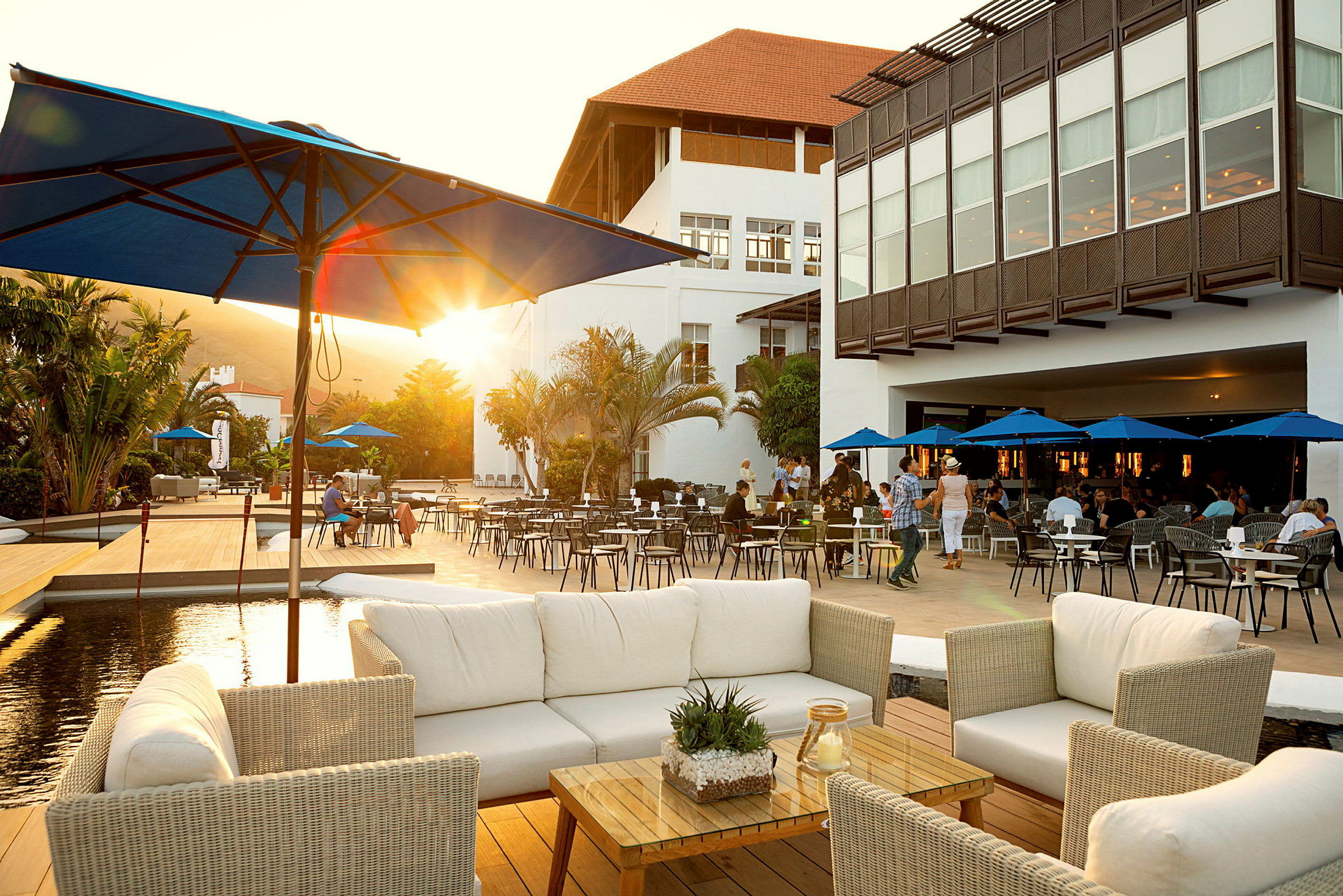 En restaurantterrasse med hvite rottingmøbler og parasoller, omgitt av et palmekledd område og en stor hvit bygningskompleks i bakgrunnen. (AI-generert tekst)