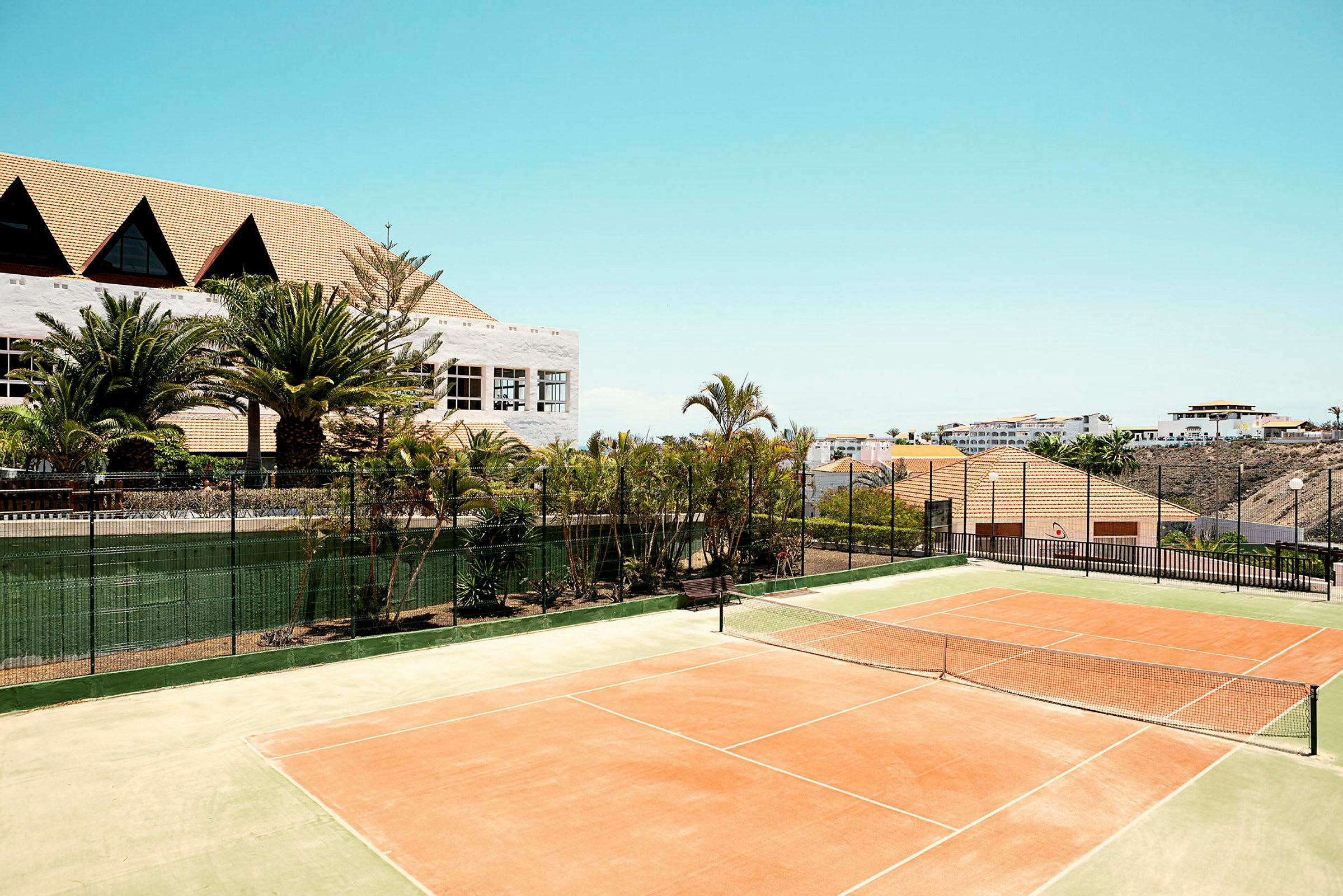 En tennisbane i solen med omgivelser av grøntområder og bygninger i bakgrunnen. (AI-generert tekst)