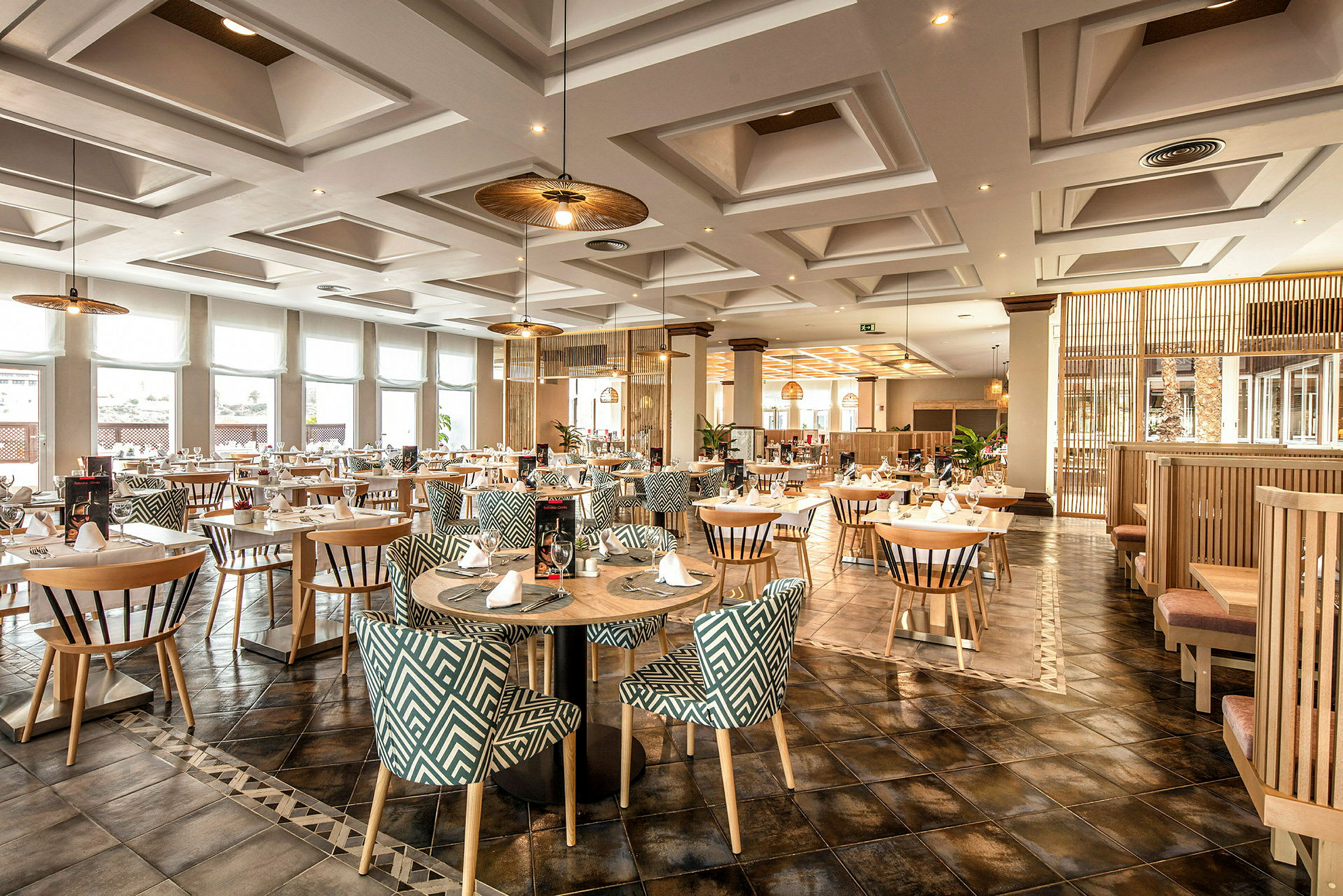 Et elegant restaurantområde med runde bord og mønstrede stoler, opplyst av taklamper og naturlig lys gjennom store vinduer. (AI-generert tekst)