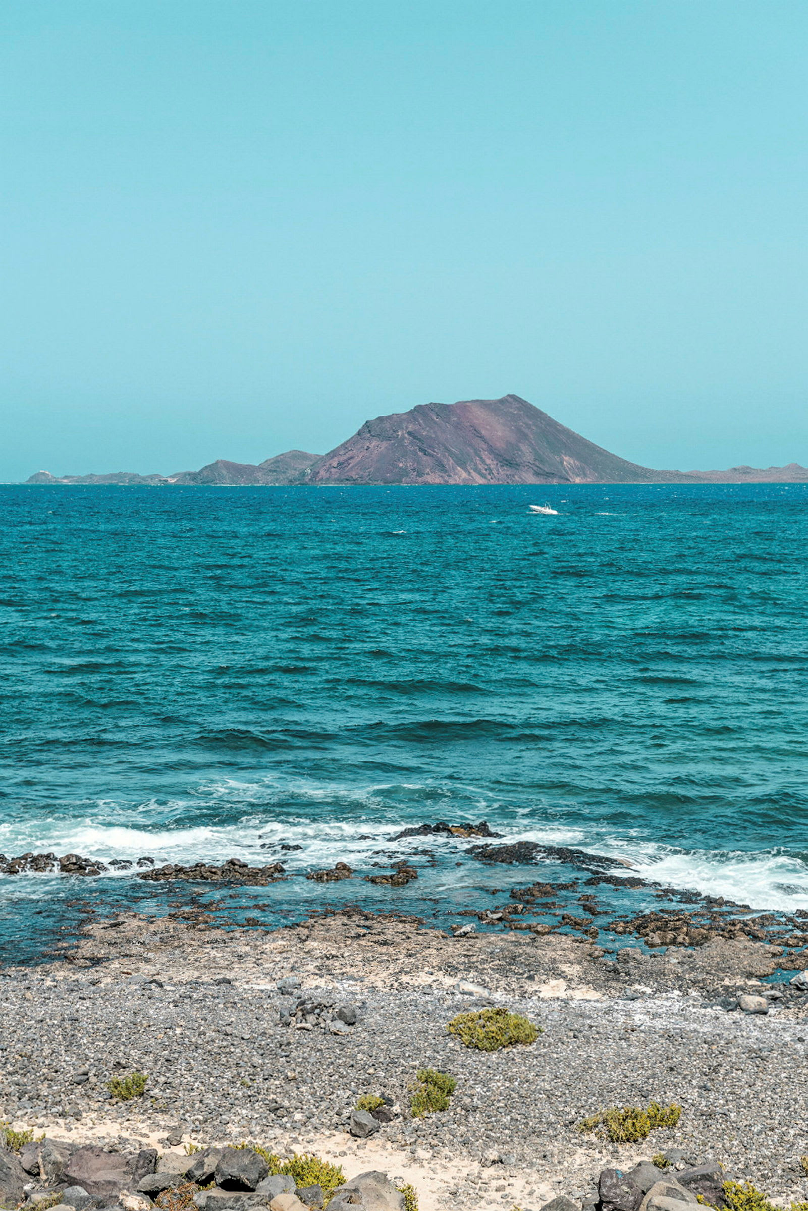 En strand med småstein og vegetasjon i forgrunnen, etterfulgt av et åpent blått hav og en vulkanlignende øy i bakgrunnen. (AI-generert tekst)
