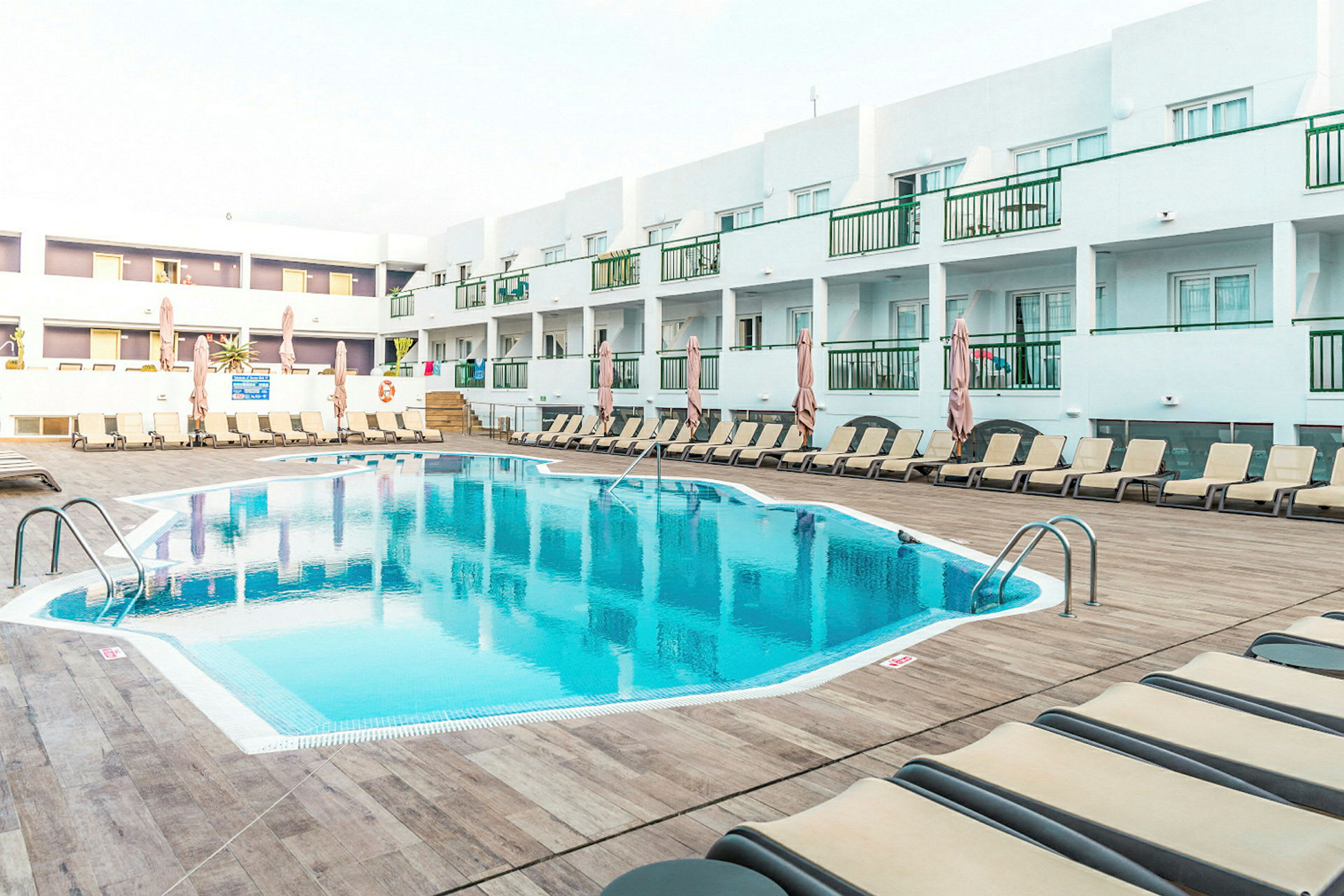 Utendørsbasseng ved et hotell, omgitt av solsenger og parasoller, med hvite bygninger i bakgrunnen. (AI-generert tekst)