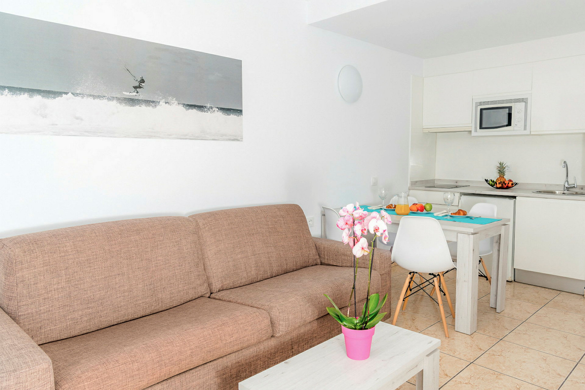 Et moderne rom med en brun sofa, et kaffebord med en rosa potteplante, et kjøkken med hvite skap og en spiseplass med et dekket bord. (AI-generert tekst)