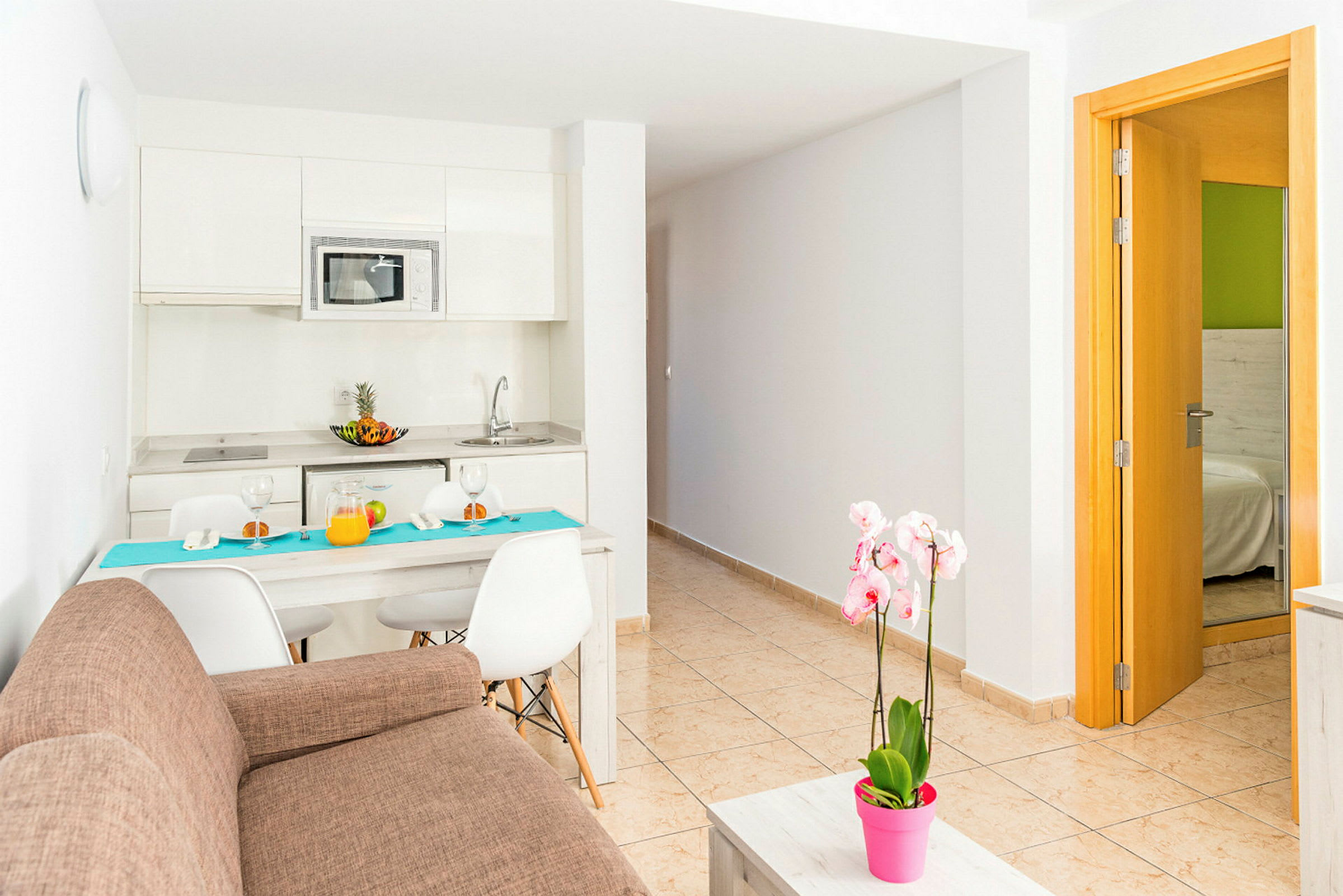 Et moderne kjøkken med spisebord og stoler, en brun sofa og en rosa blomsterpotte på et sofabord. Inngang til soverommet er synlig til høyre. (AI-generert tekst)