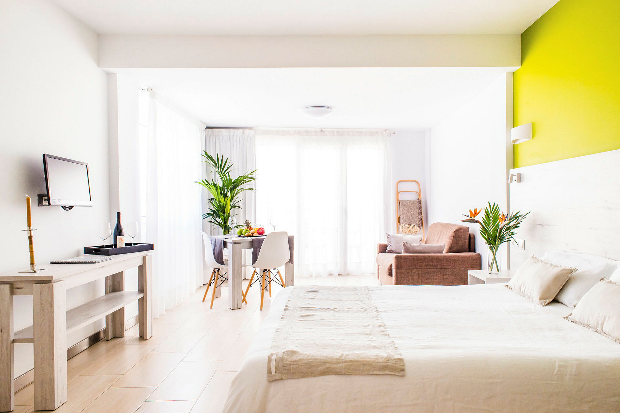 Et moderne innredet hotellrom med en seng, sittegruppe og spisebord, dekorative planter og store vinduer. (AI-generert tekst)