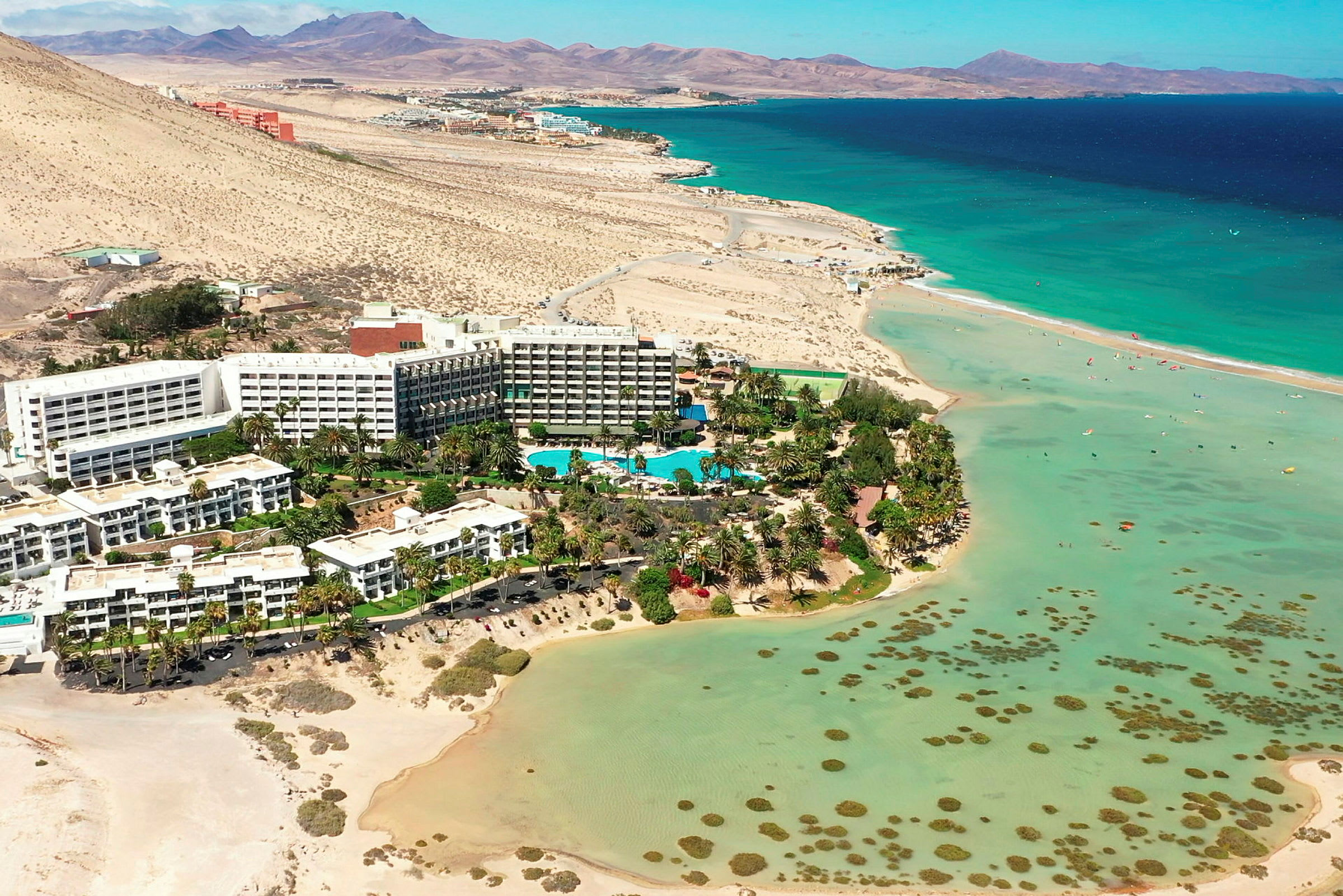 Hotellkompleks ved en strand med turkist vann og omkringliggende ørkenlandskap. (AI-generert tekst)