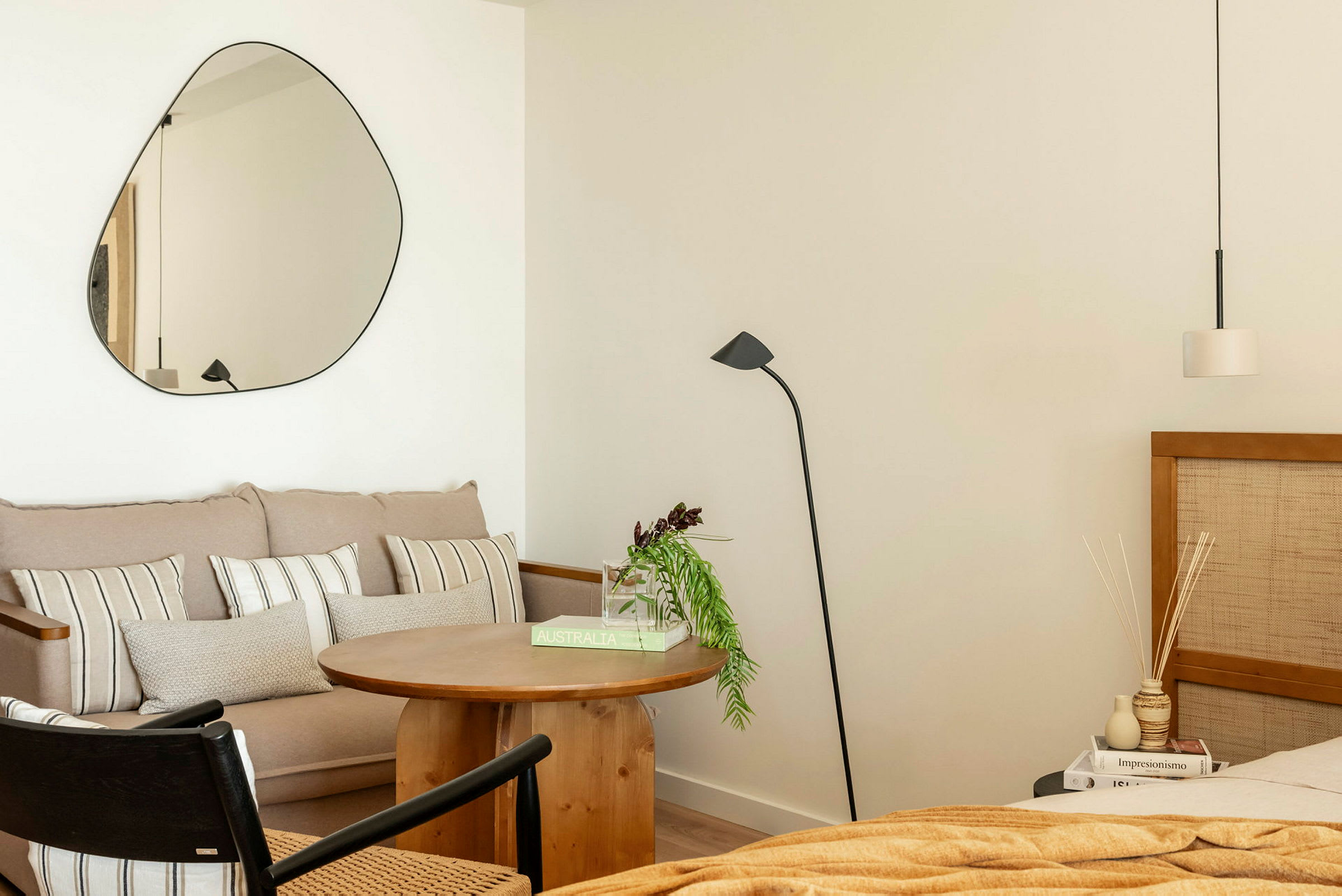 Et rom med moderne interiør. En sofa med puter, et rundt trebord, en gulvlampe og et speil på veggen. Til høyre sees en seng og et nattbord. (AI-generert tekst)