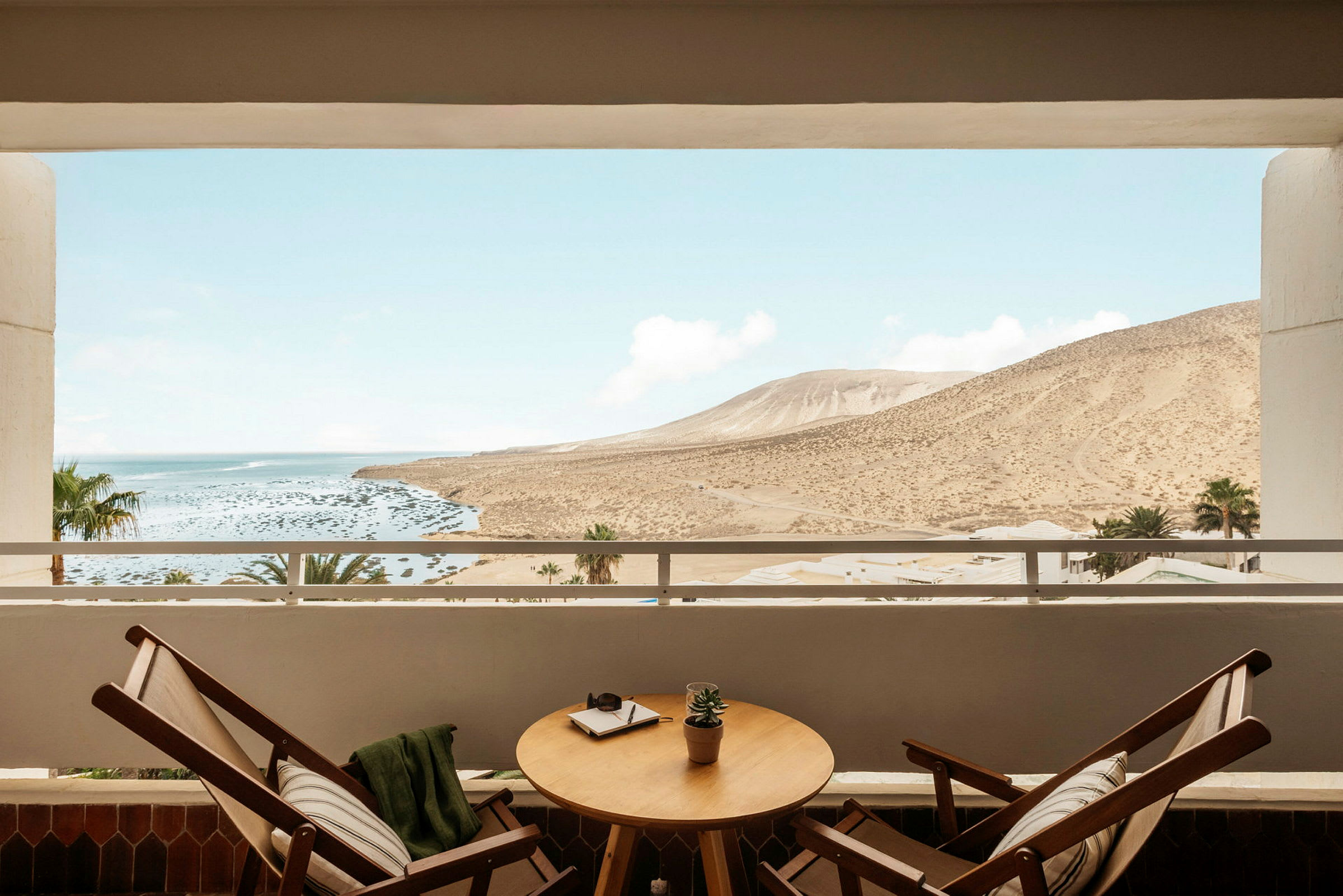 En balkong med to stoler og et bord, utsikt over et landskap med fjell og kyst. (AI-generert tekst)