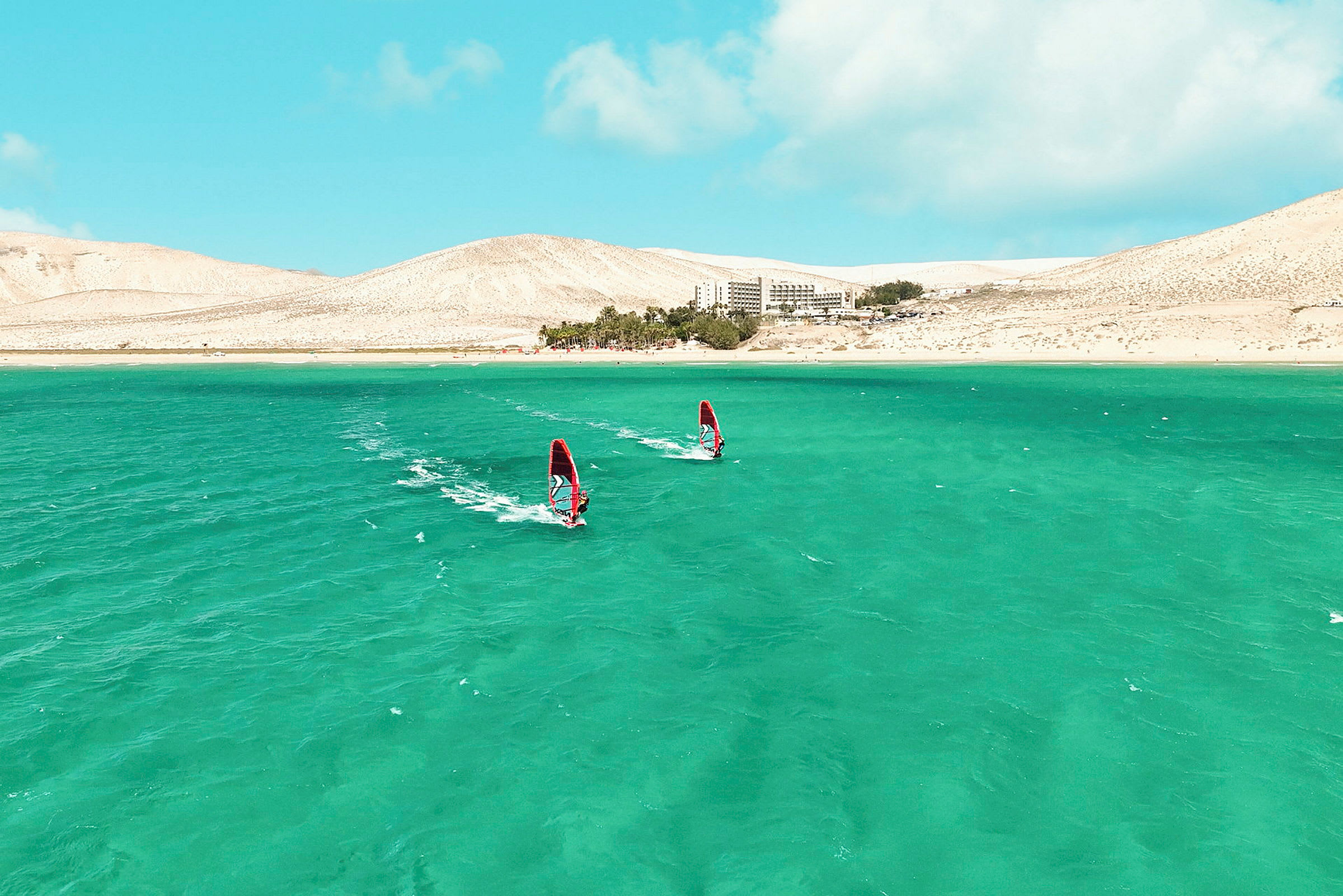 To windsurfere på blågrønt vann med et sandete landskap og en bygning i bakgrunnen. (AI-generert tekst)
