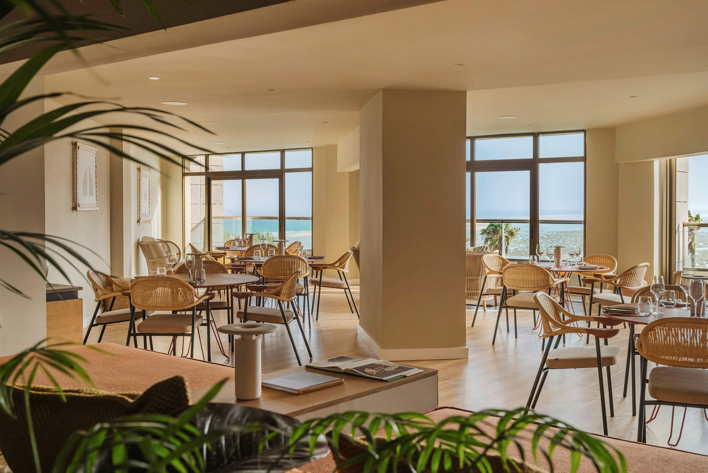 En luftig restaurant med store vinduer som gir utsikt mot havet. Det er flere bord med stoler, og interiøret er lyst og elegant. (AI-generert tekst)