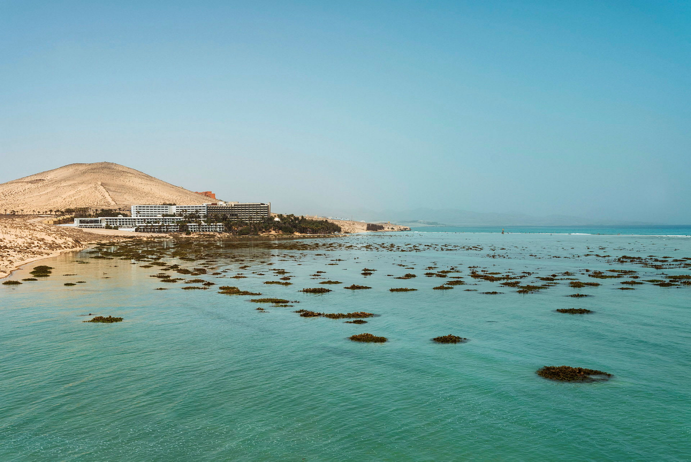 Hotellbygninger nær en strand med klart blått vann og spredte klynger av vegetasjon. (AI-generert tekst)