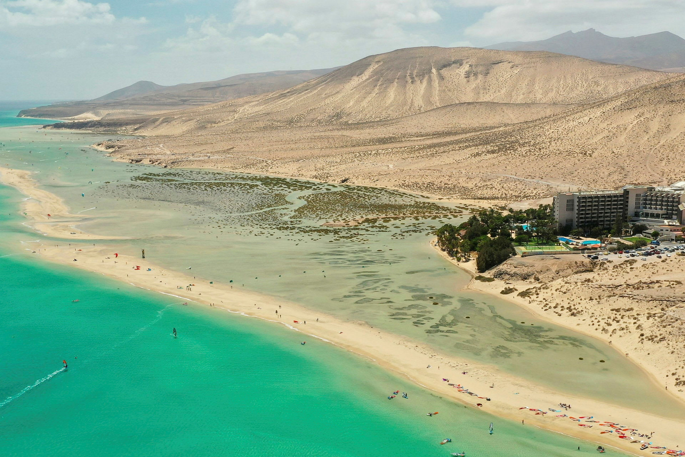 Strand med klart vann og sanddyner i bakgrunnen. Hotellbygning og trær vises på den ene siden. (AI-generert tekst)