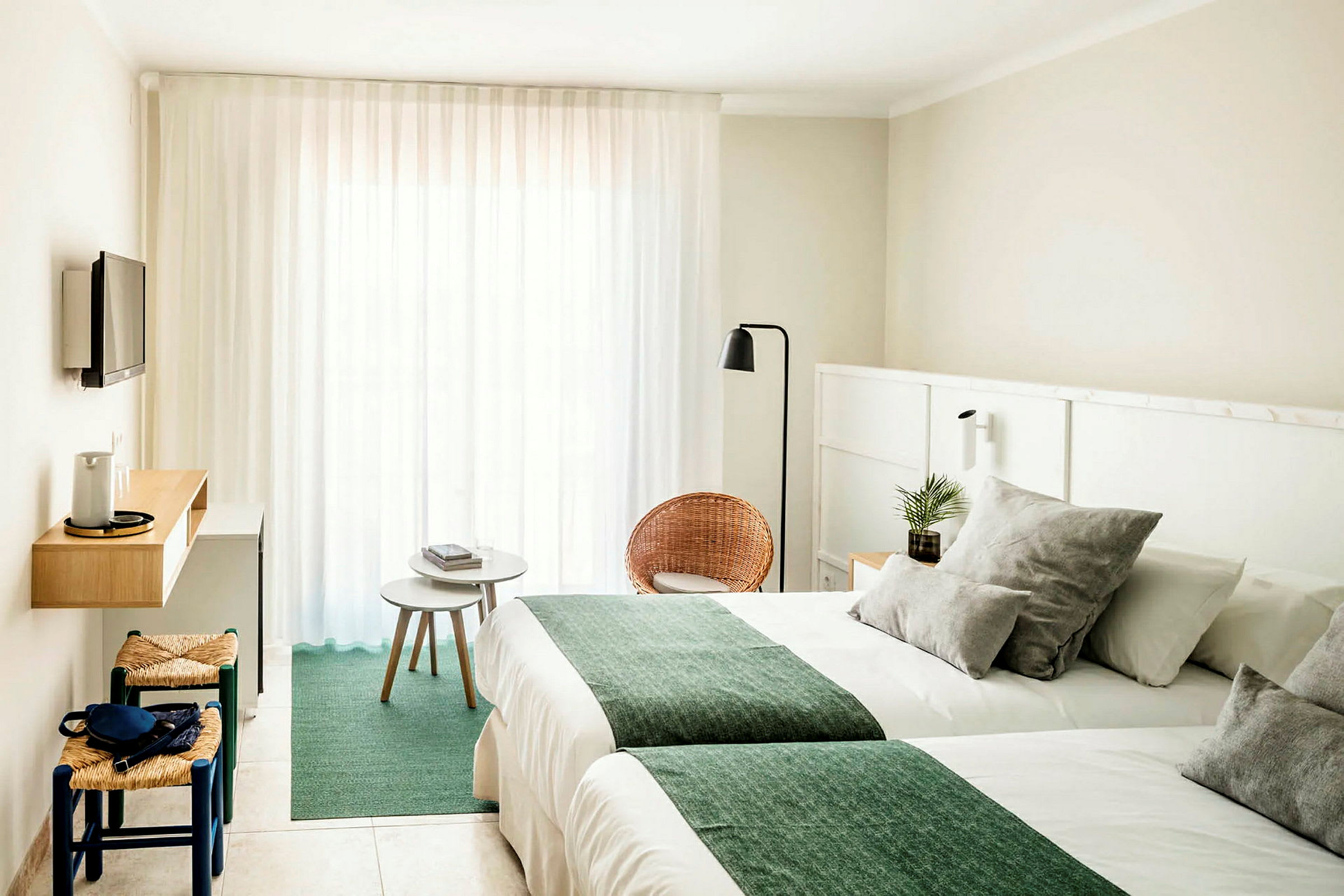 Et hotellrom med lyse vegger, to senger med grønne tepper, en liten sittegruppe med stoler og bord, samt en veggmontert TV. (AI-generert tekst)