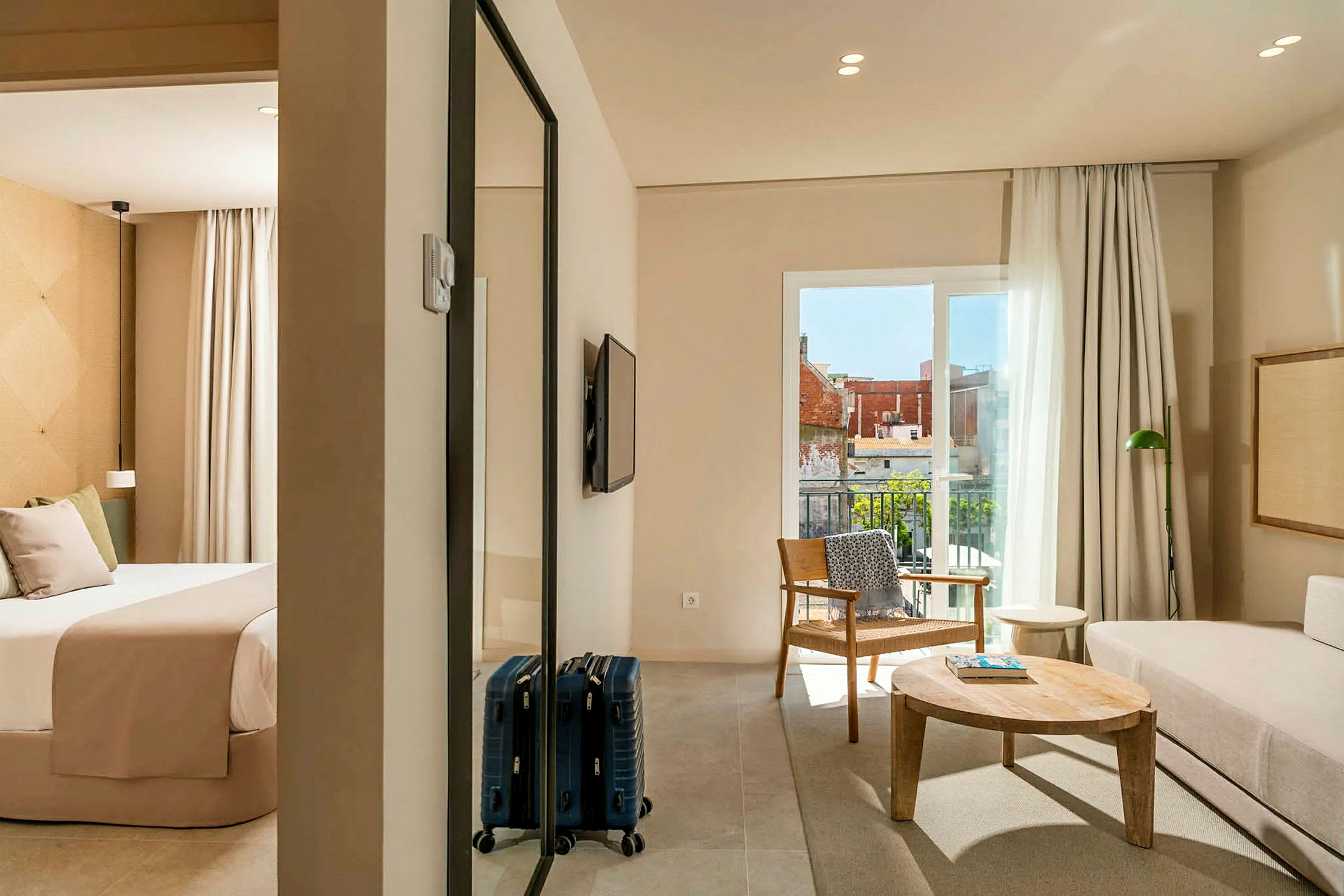 Et hotellrom med et soverom og stue. Soverommet har en seng med beige sengetøy, og stuen har en sofa, et trebord og en lenestol. Det er en balkong med utsikt. (AI-generert tekst)