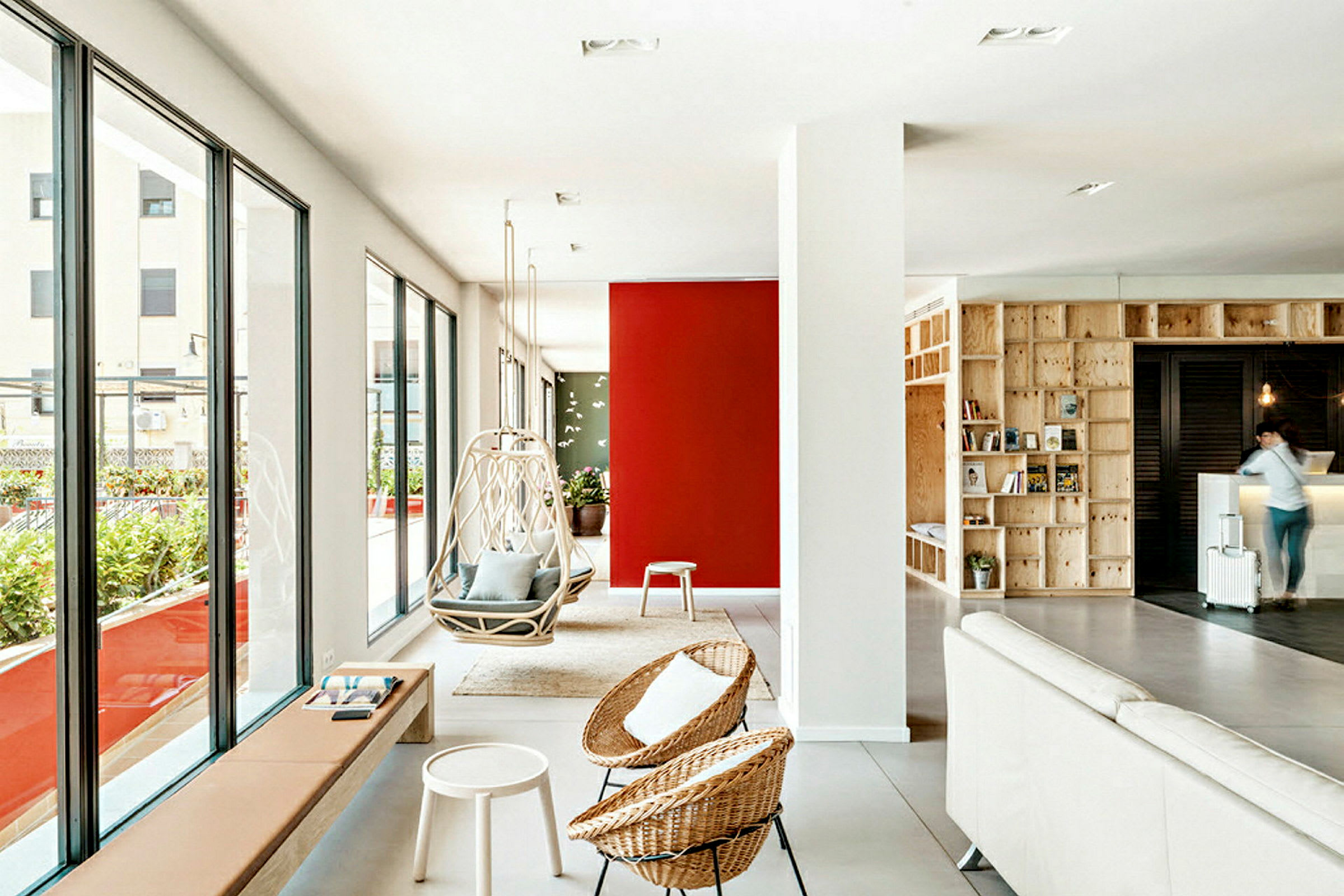 Et moderne og lyst inneområde med store vinduer som vender ut mot en terrasse. Rødt og mønstret på veggen gir rommet farge. Hengestoler står ved siden av andre designmøbler og detaljer. (AI-generert tekst)