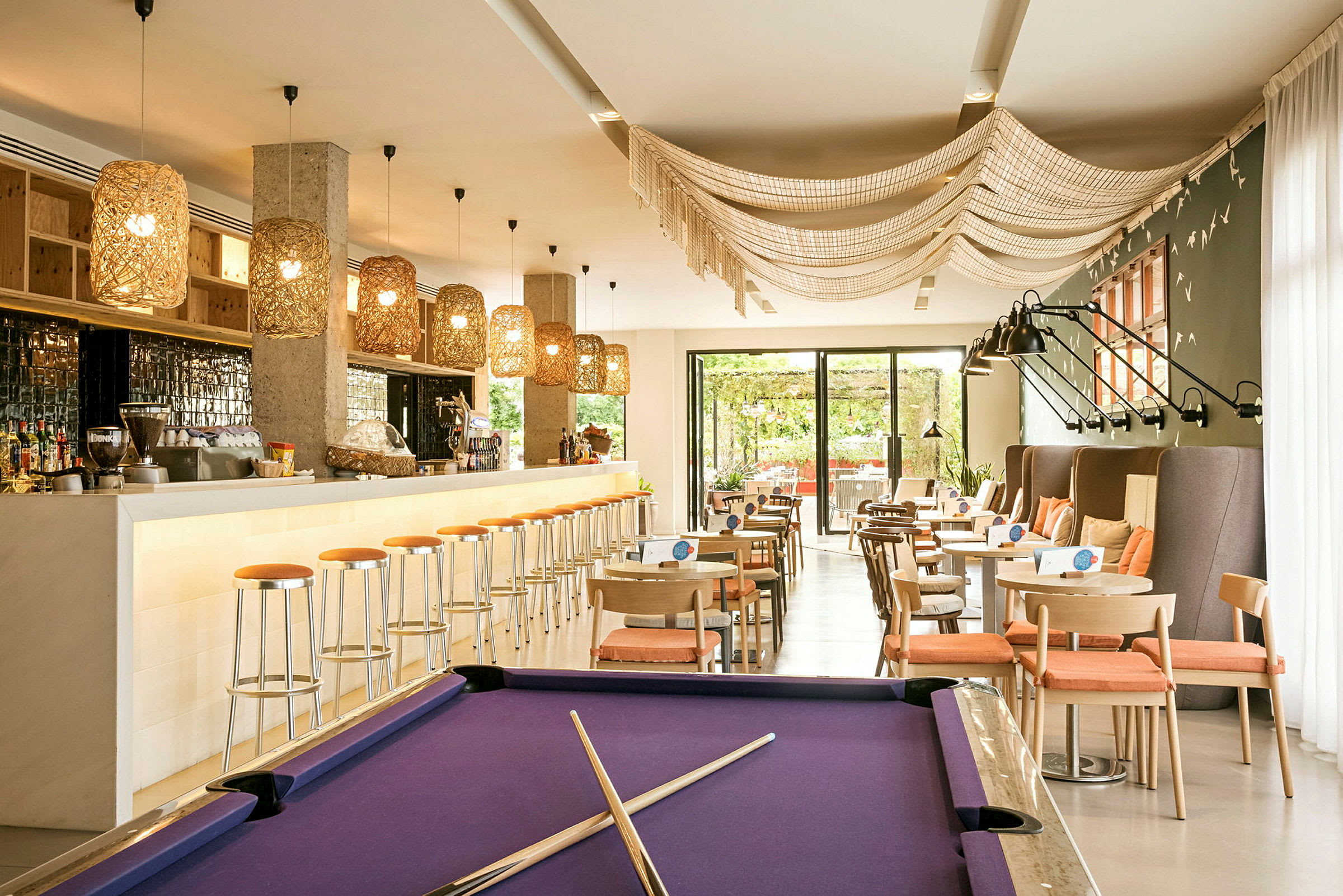 Et moderne og lyst rom med en bar med høye stoler, bord med oransje sitteputer og et biljardbord med lilla duk. (AI-generert tekst)