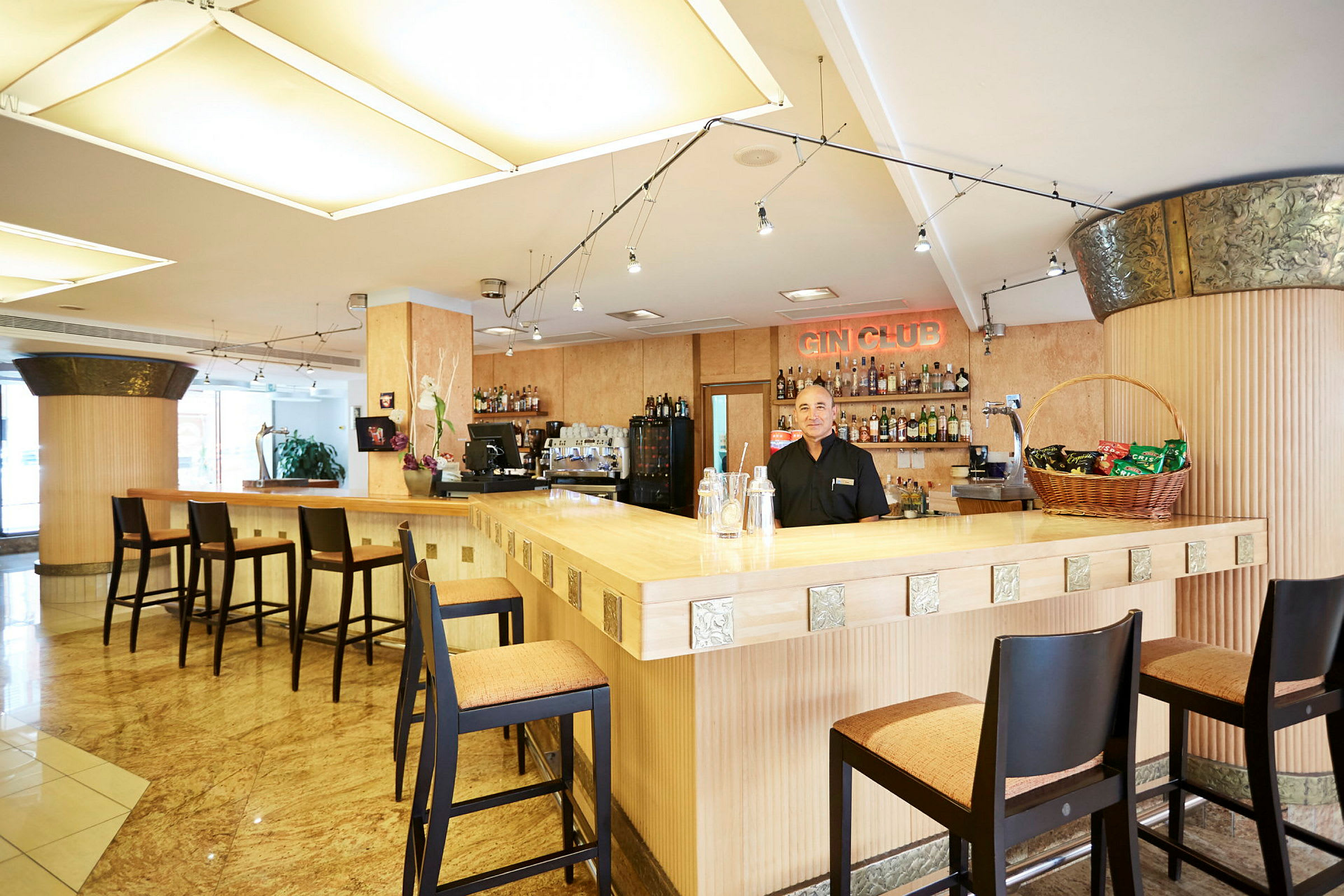 En lys barinteriør med høye stoler rundt en tremonter. På bakveggen sees flere flasker og et skilt med teksten "Gin Club". (AI-generert tekst)