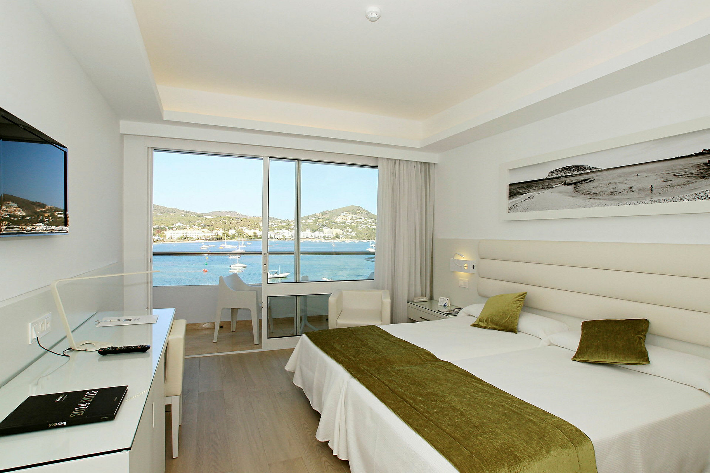 Et lyst og moderne hotellrom med to senger og store glassdører som fører ut til en balkong. Utenfor er det utsikt over havet og det omkringliggende landskapet. (AI-generert tekst)