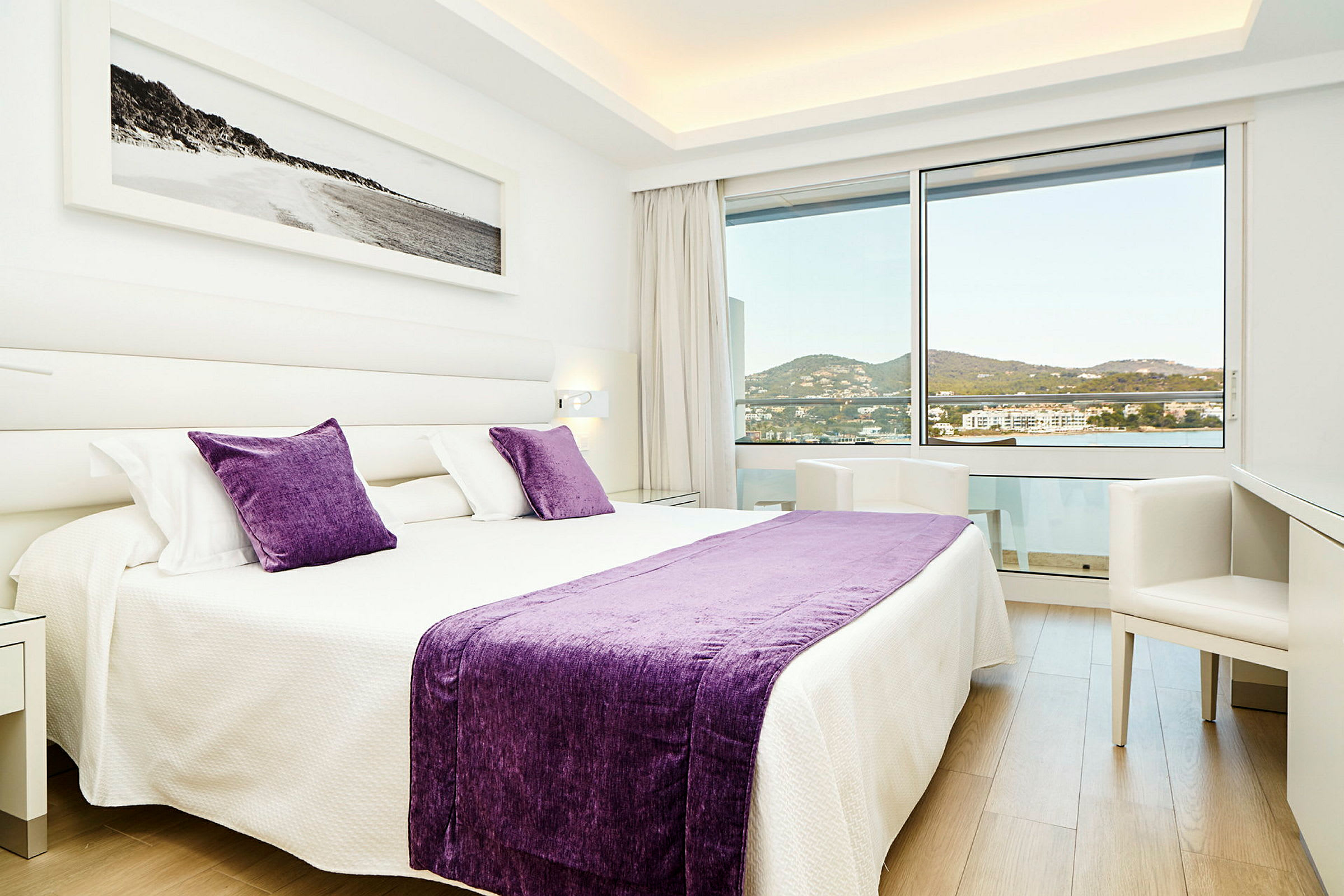 Et moderne hotellrom med en stor seng dekorert med lilla puter og teppe, lyse vegger og et stort vindu som gir utsikt over området. (AI-generert tekst)