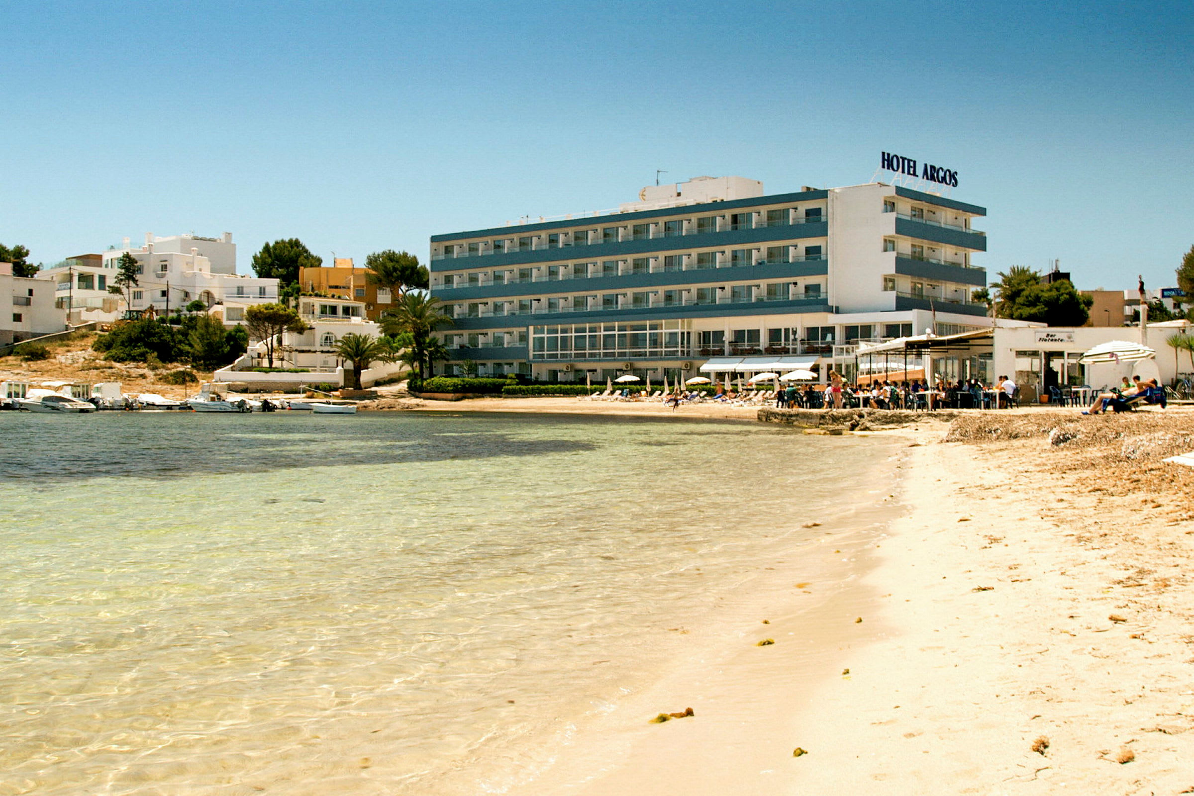 Strand med klart vann foran et hotell og bygninger. (AI-generert tekst)