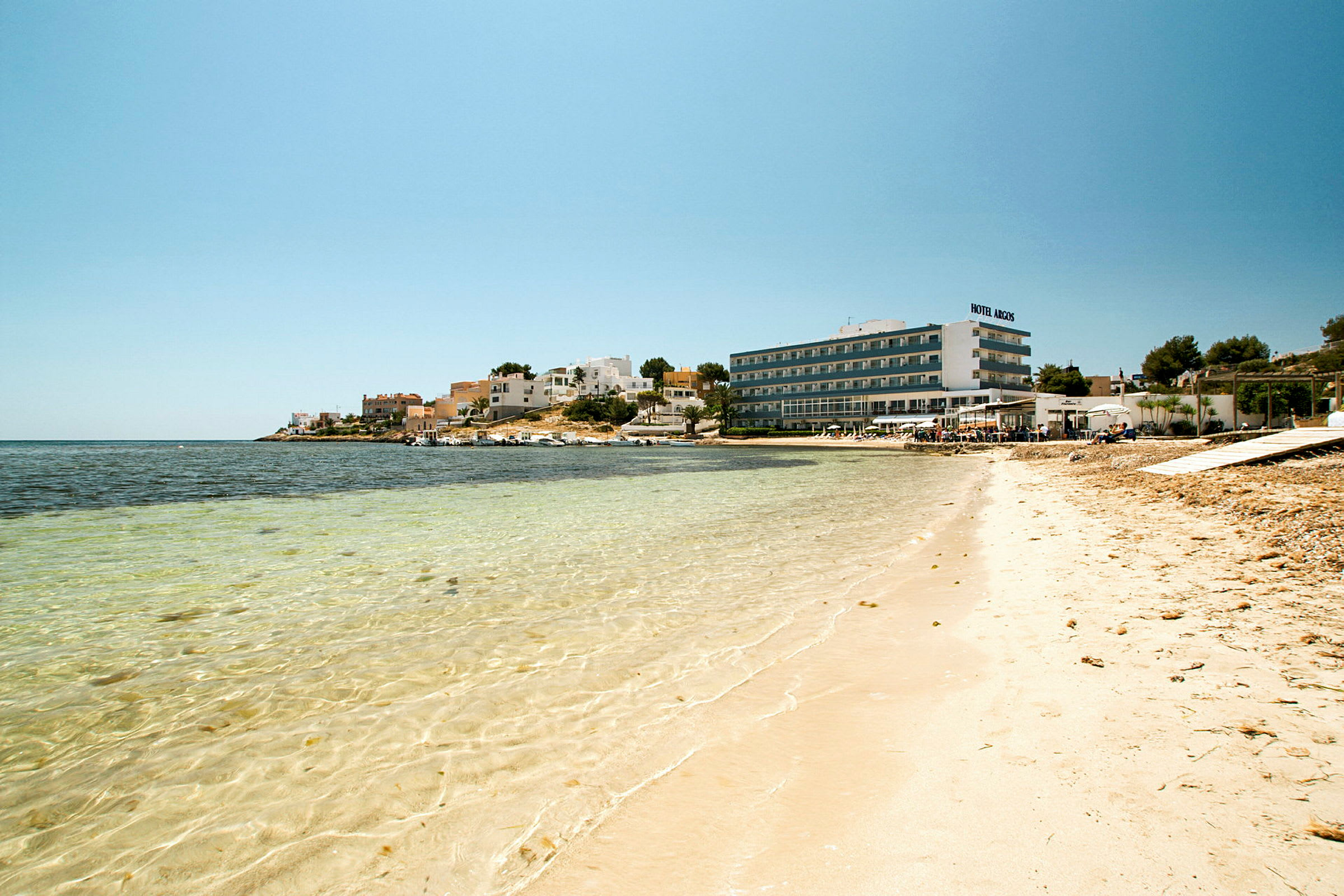 En strand med klart vann, bygninger og et hotell i bakgrunnen. (AI-generert tekst)