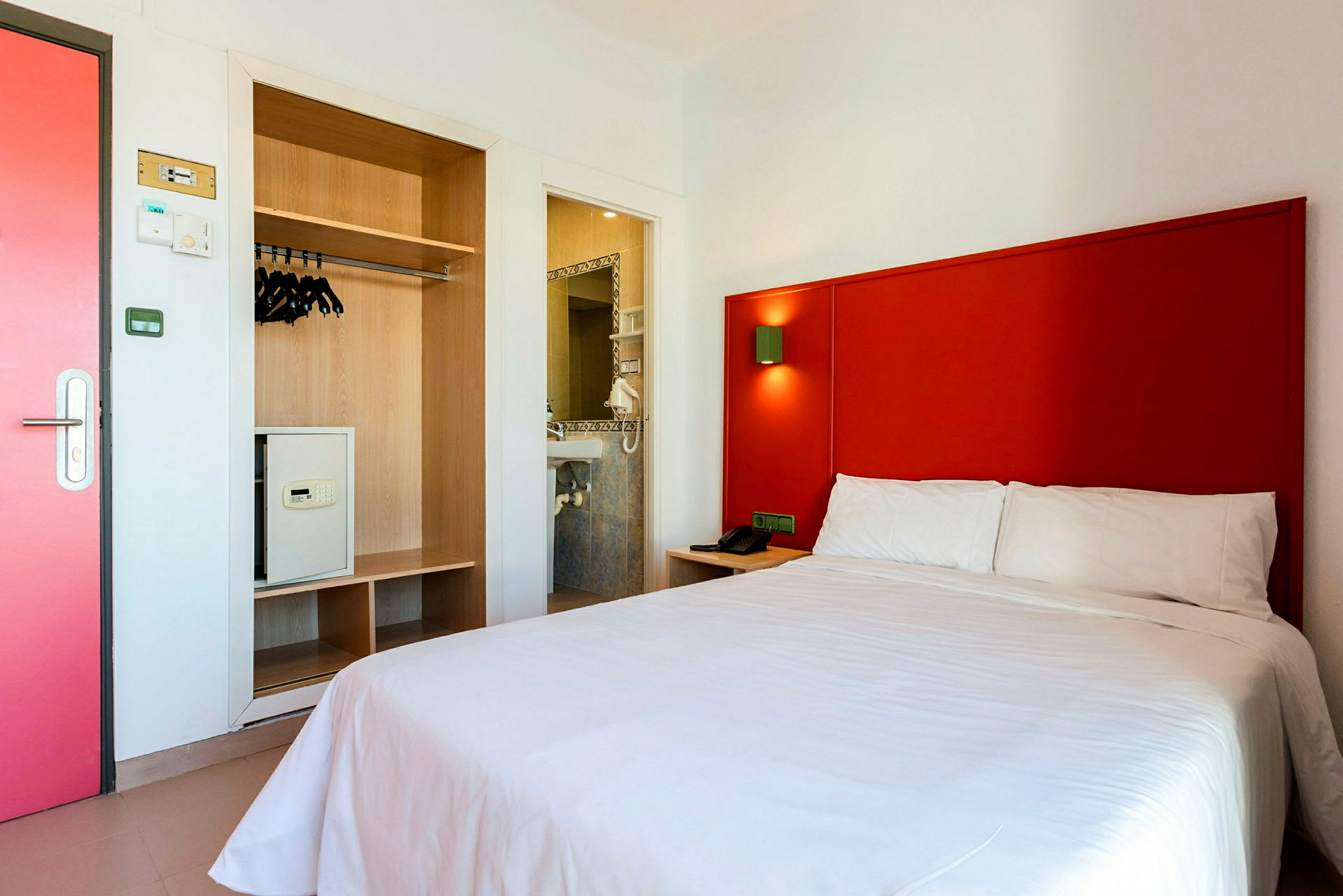Hotellrom med rødt sengegavl, hvit seng, og garderobe med safe. (AI-generert tekst)