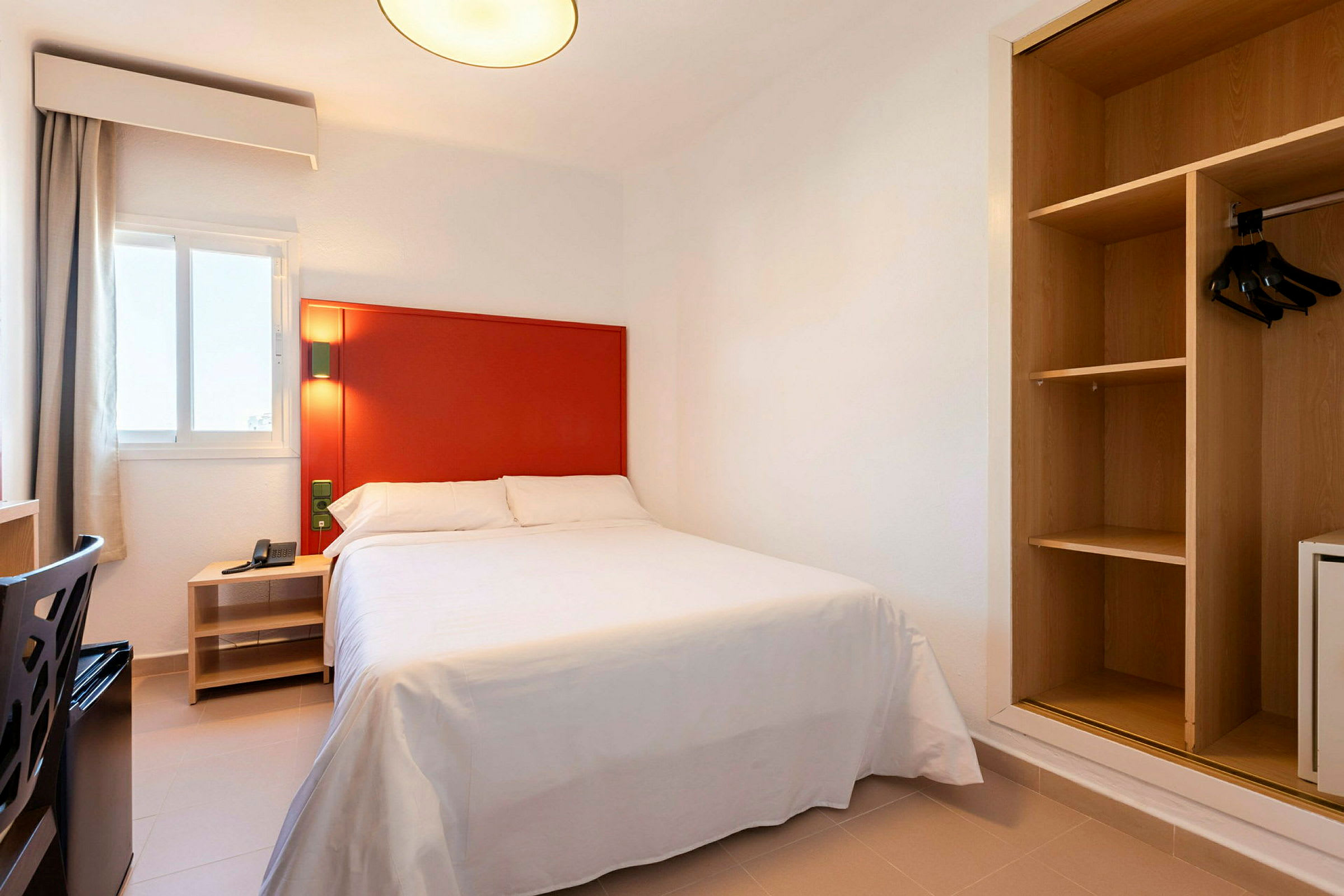 Bilde av et hotellrom med en seng, rød sengegavl og åpen garderobe. (AI-generert tekst)