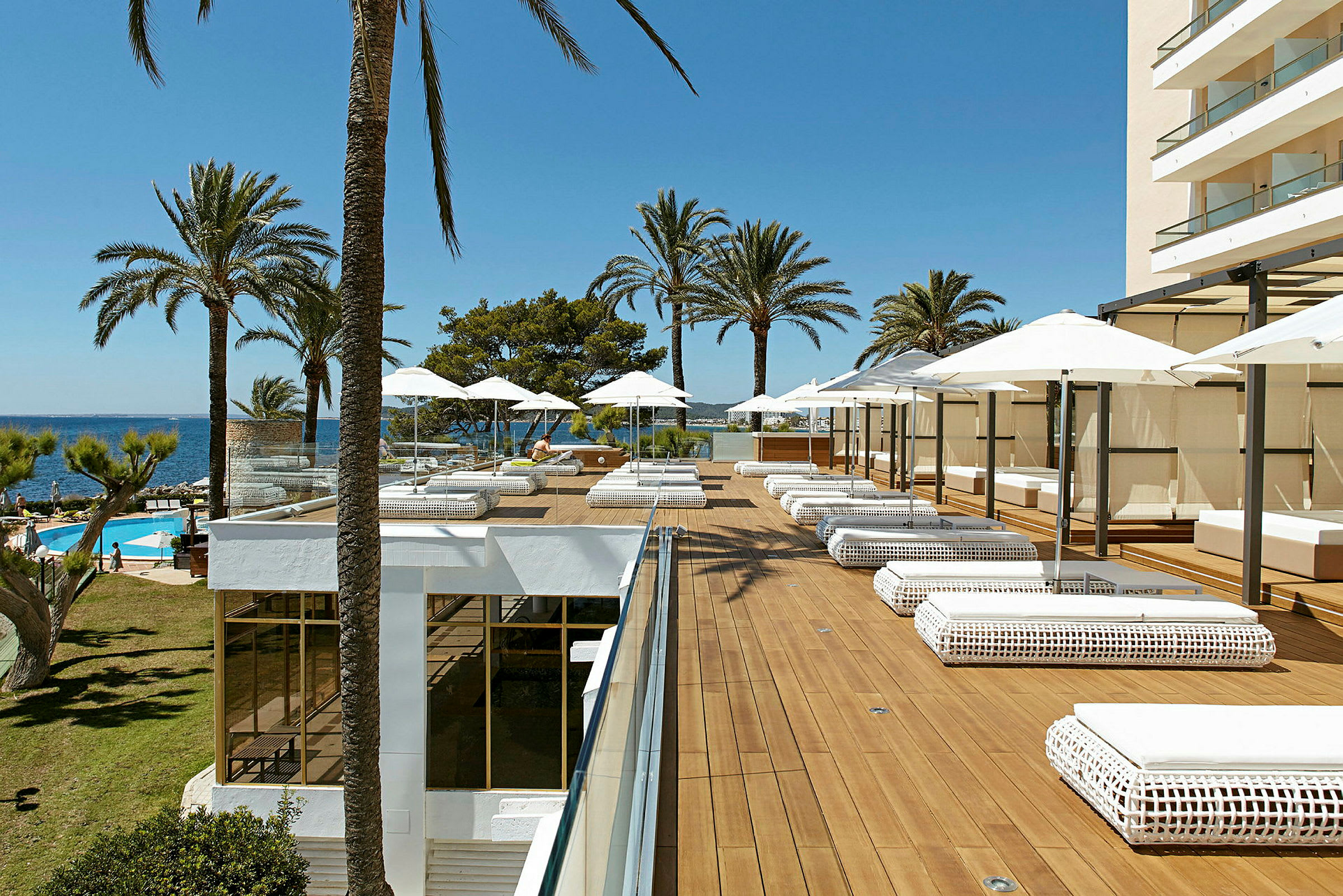 En hotellterrasse med solsenger og parasoller, omgitt av palmer og utsikt mot havet. (AI-generert tekst)