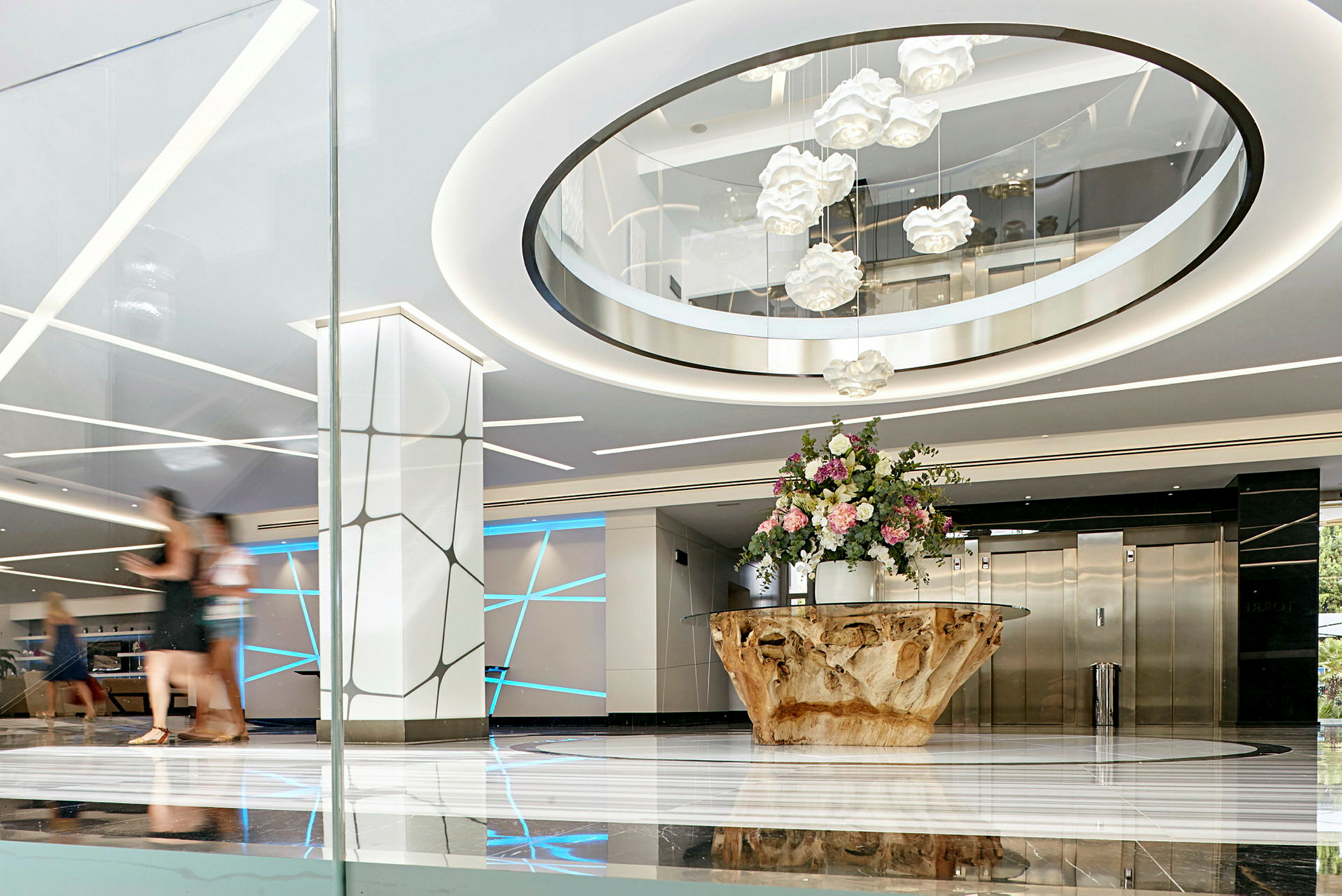 Lys og moderne hotellobby med et stort bord av tre og en bukett blomster i midten, elegante lyseffekter og heiser i bakgrunnen. (AI-generert tekst)