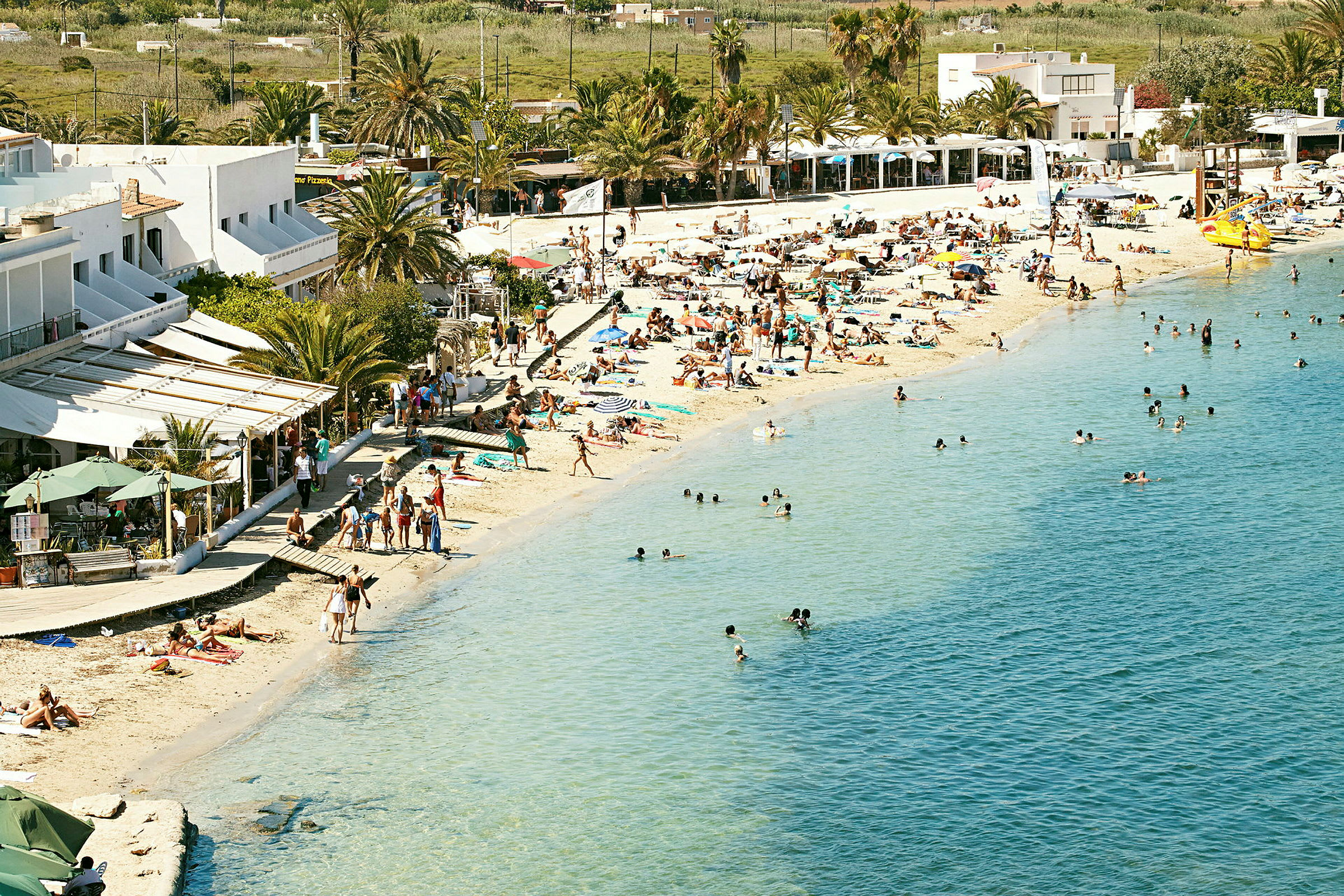 Strandliv med mennesker som soler seg på stranden og svømmer i havet, bygninger og palmer i bakgrunnen. (AI-generert tekst)
