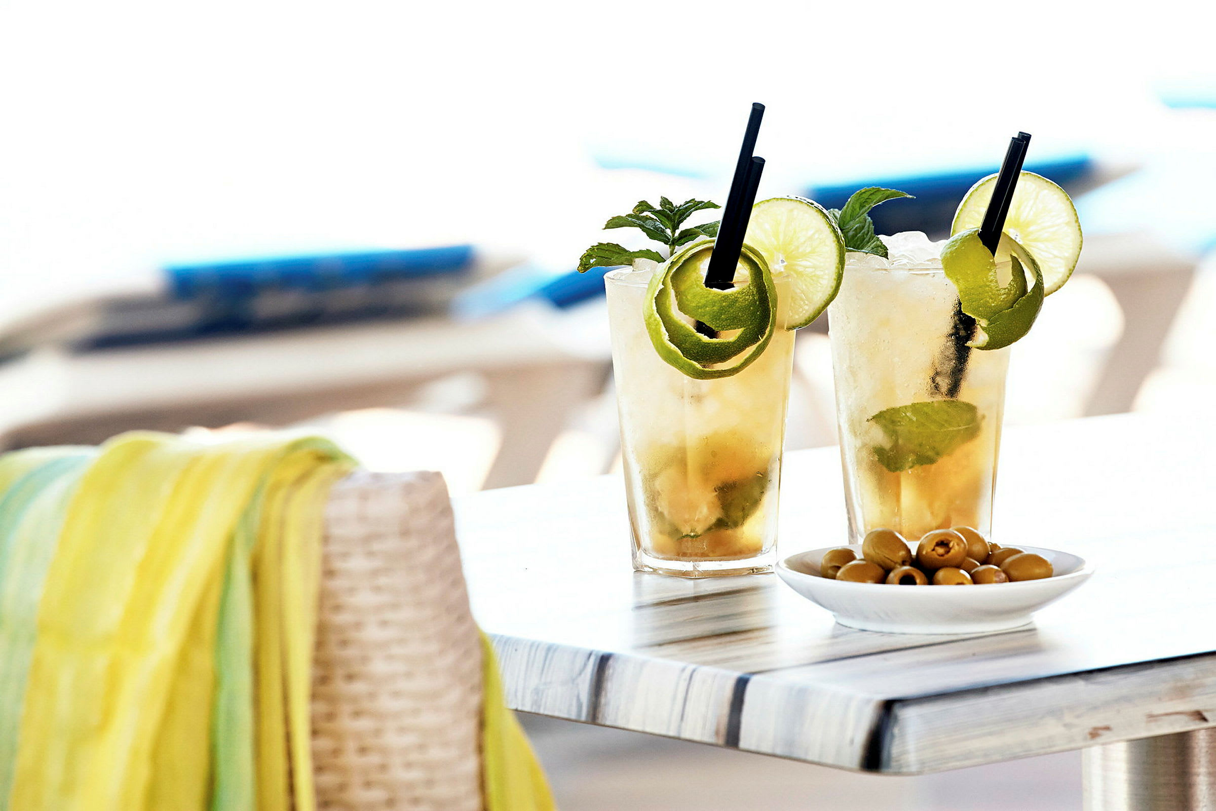 To glass med kjølte cocktails dekorert med lime og mynte står på et bord ved siden av en skål med oliven. (AI-generert tekst)