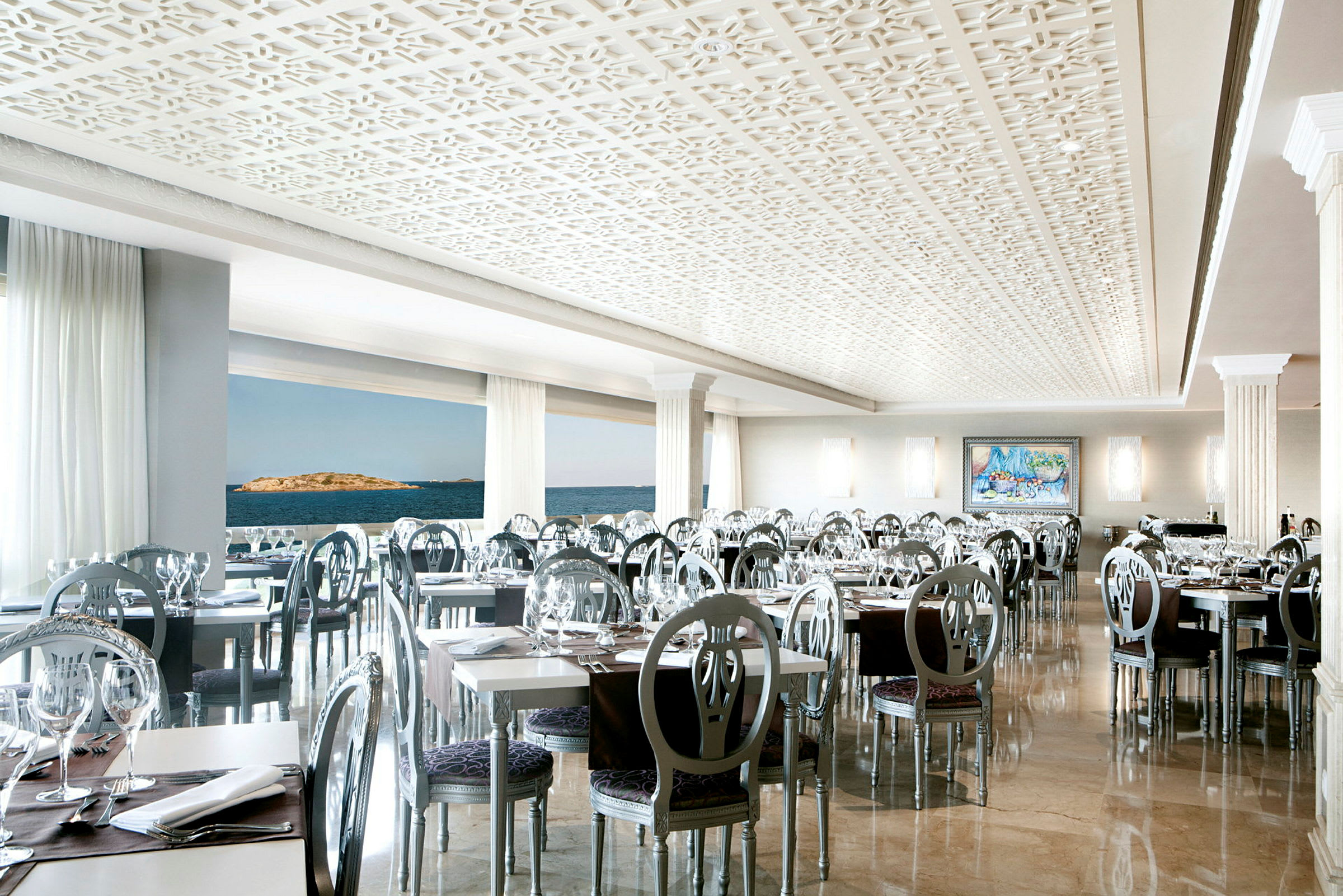 En elegant innredet restaurant med moderne stoler og hvite bord, med store vinduer som viser utsikt over havet. (AI-generert tekst)