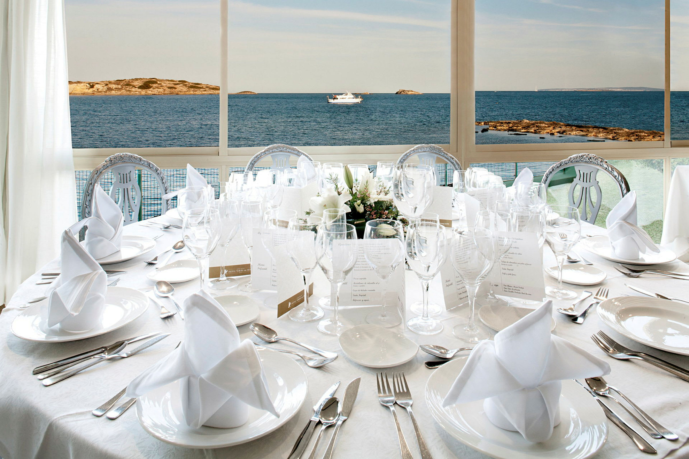 Bord dekket med hvitt servise, elegant bestikk og glass foran et vindu med utsikt over havet og kysten. (AI-generert tekst)