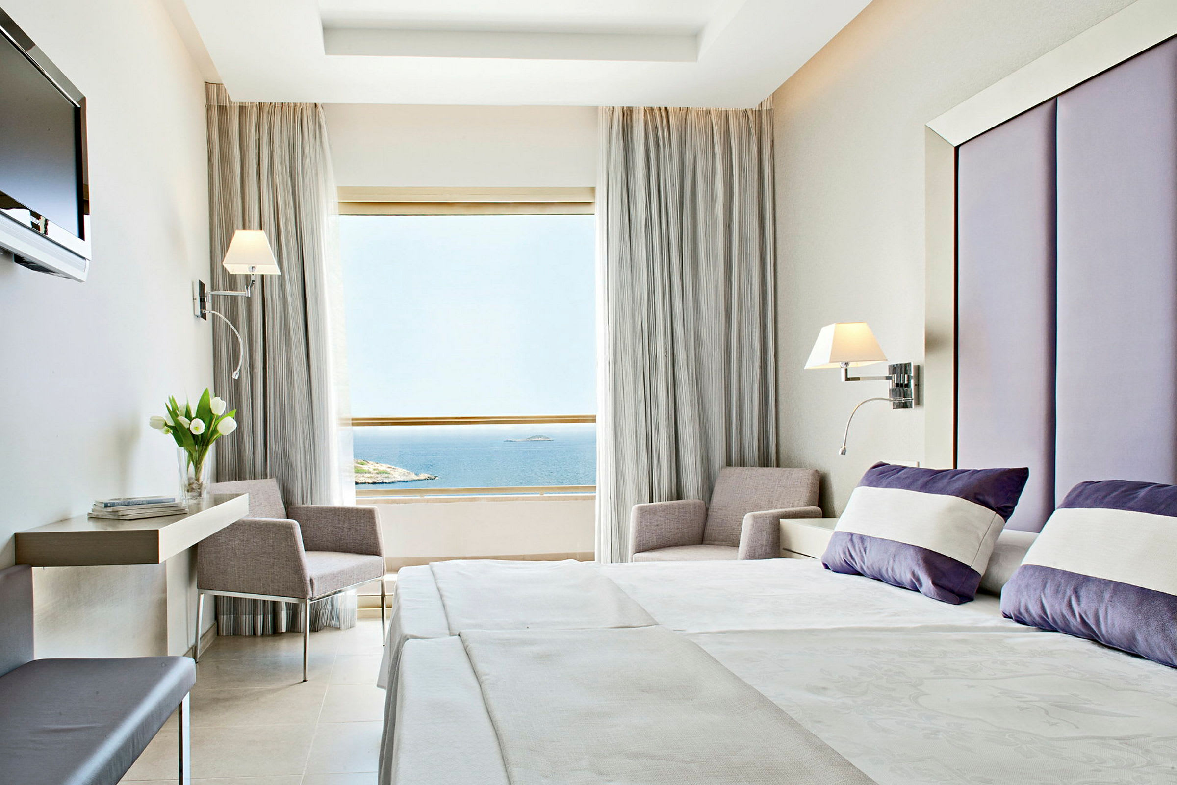 Et hotellrom med moderne innredning. Sengen er oppredd med hvite og lilla puter, og et stort vindu med utsikt over havet er synlig. (AI-generert tekst)