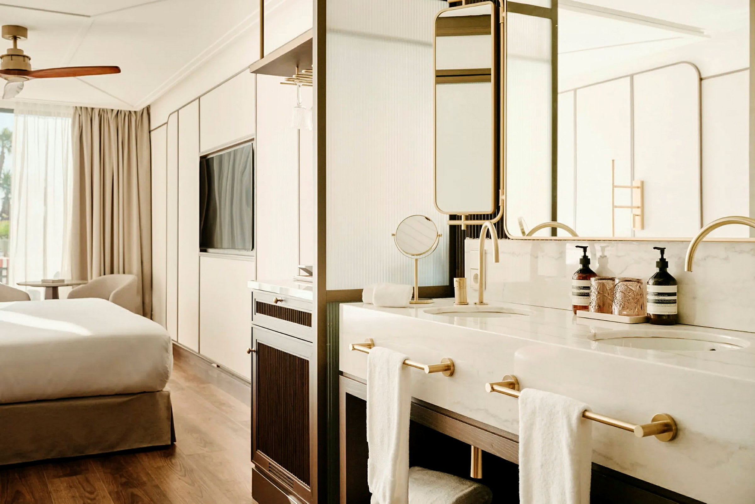 Et elegant hotellrom med en sovedel og et åpent bad med to servantområder og gullfargede armaturer. (AI-generert tekst)