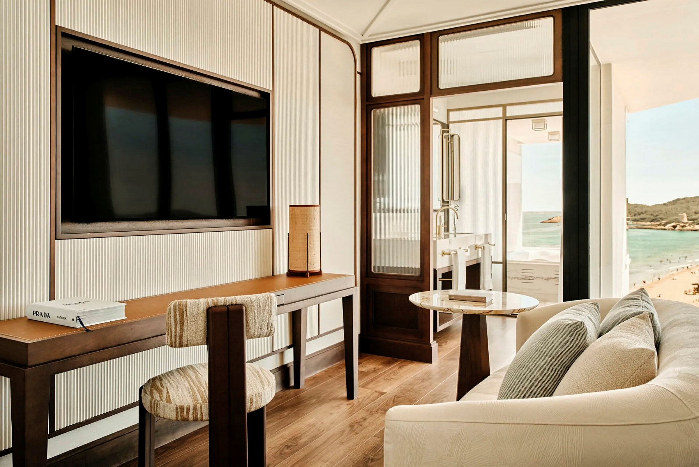 Et moderne hotellrom med skrivebord, stol, sofa og utsikt over stranden og havet fra vinduet. (AI-generert tekst)