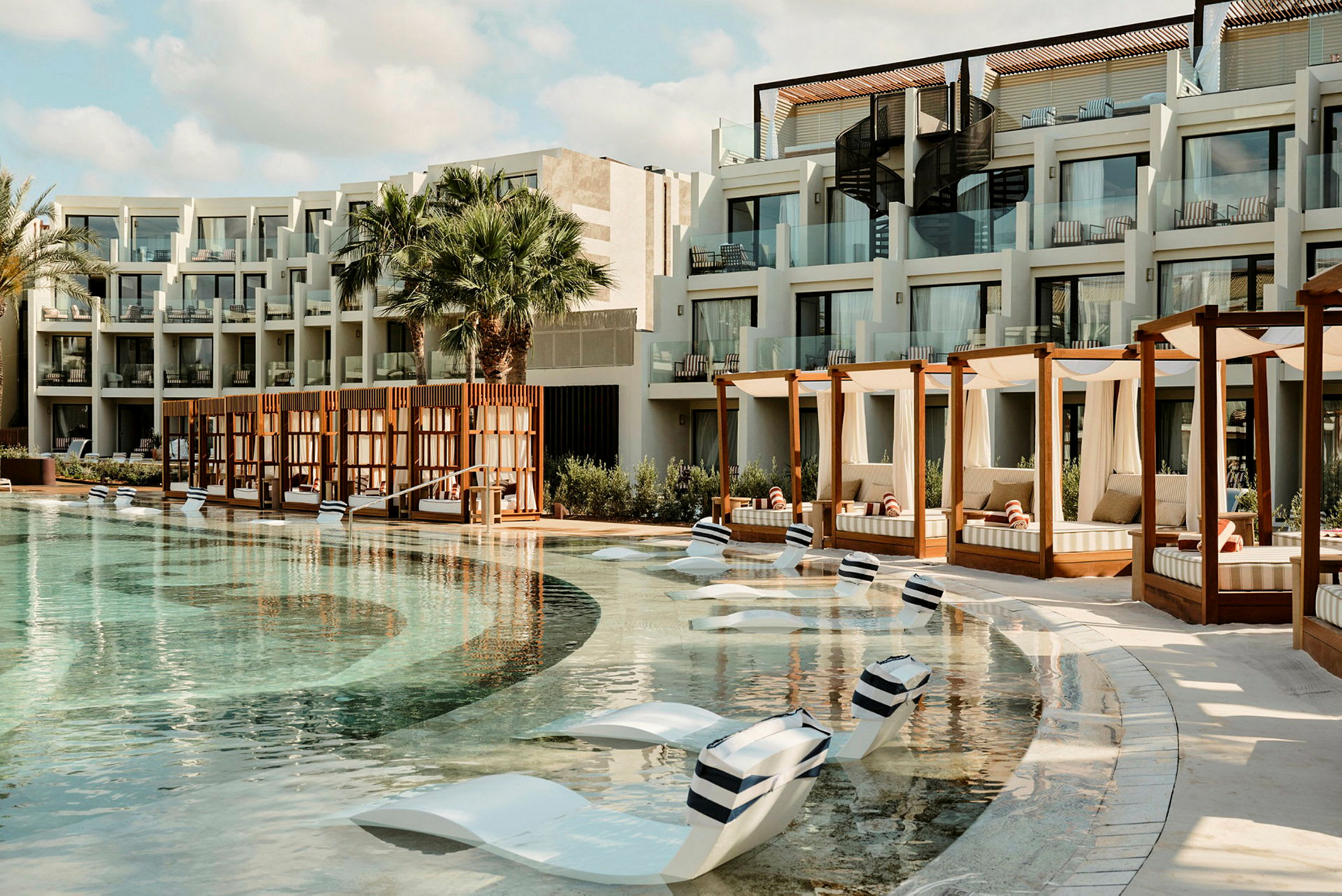 Et hotellbasseng med solsenger i bassenget og cabanas ved siden. Bygningen har balkonger med glassrekkverk. (AI-generert tekst)