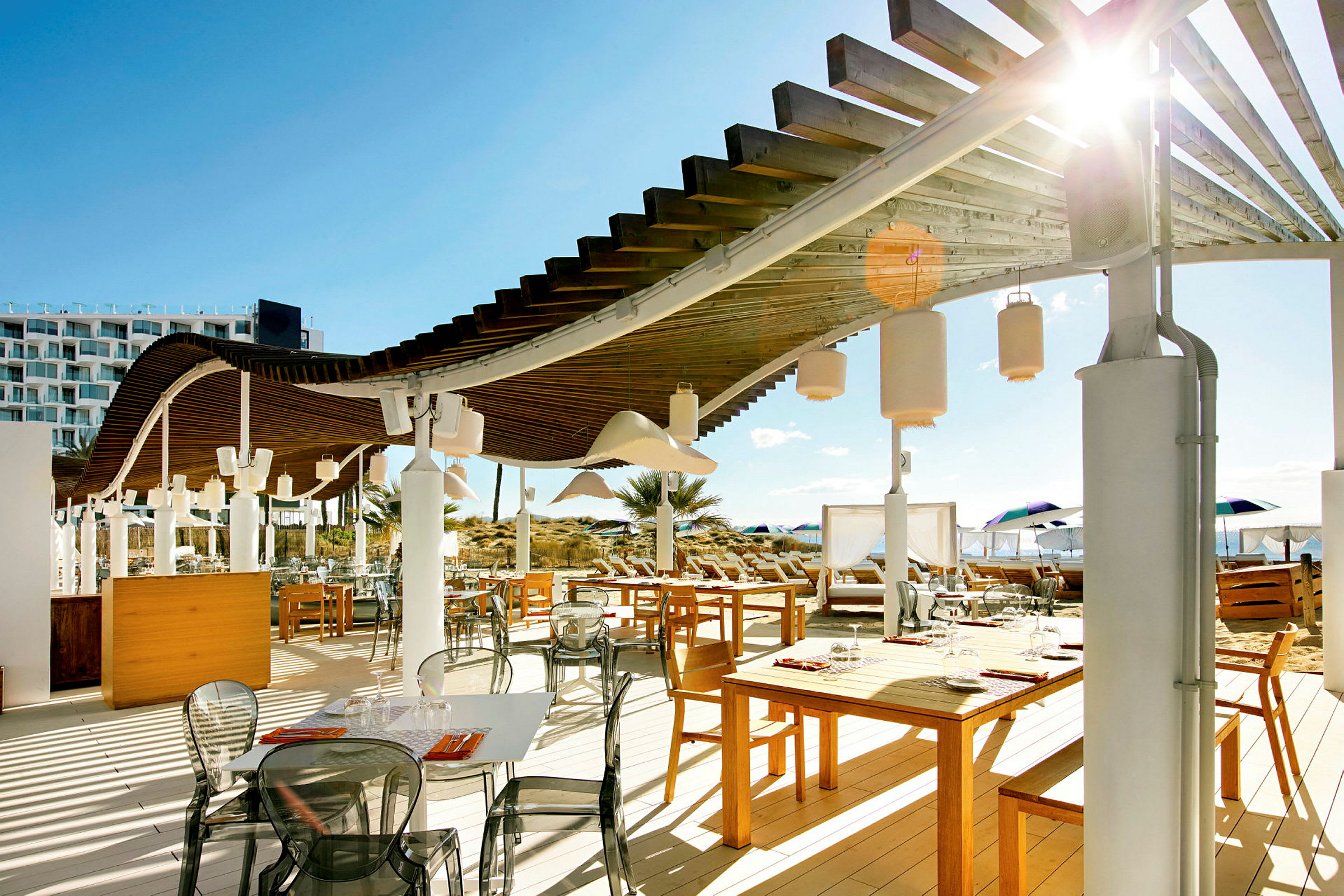 En utendørsrestaurant med trebord, stoler og en pergola over. I bakgrunnen sees strand og parasoller. (AI-generert tekst)