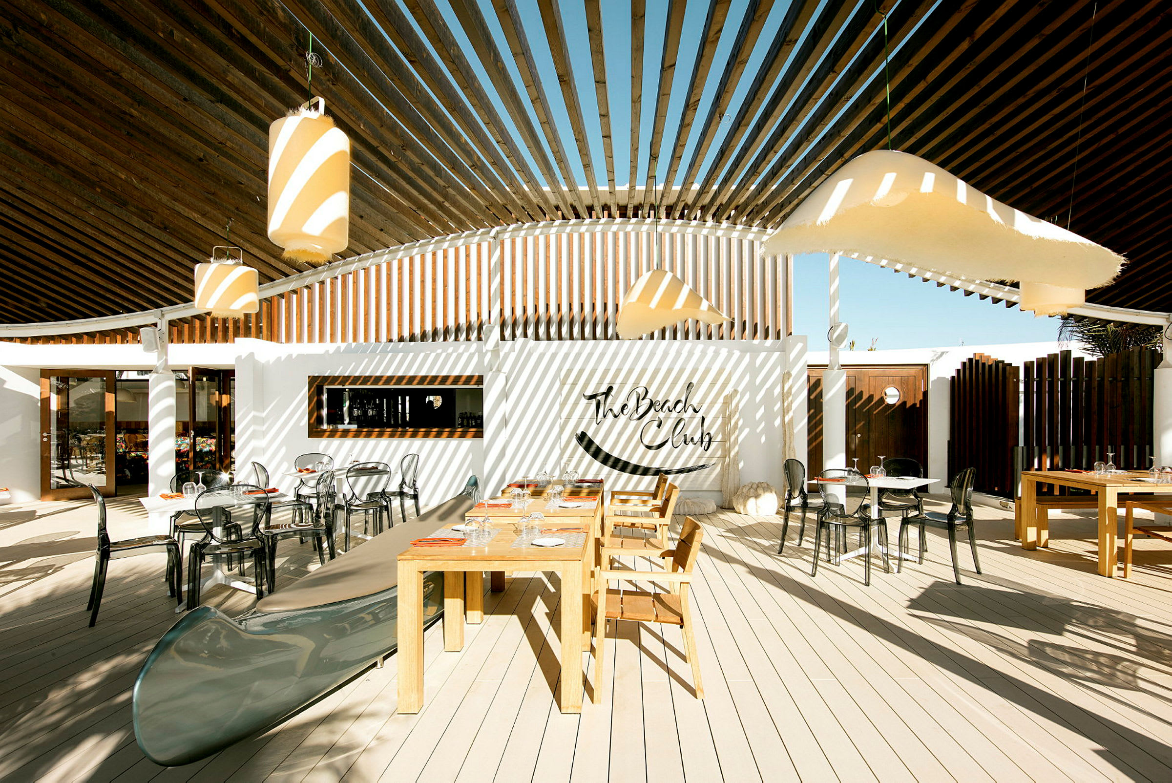Utendørsområde med bord og stoler, samt dekorasjon med The Beach Club-skiltet. (AI-generert tekst)