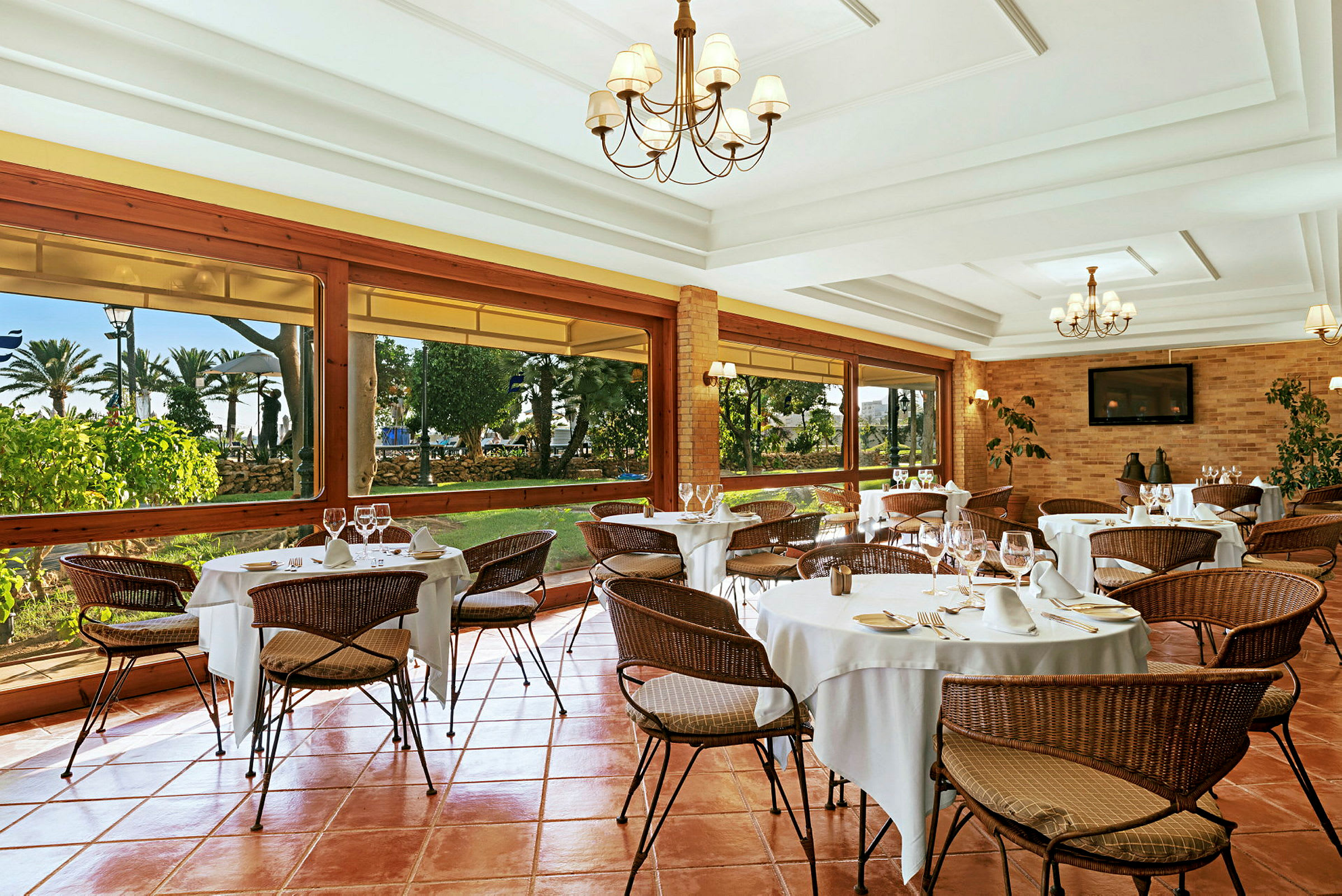 En elegant restaurant med dekkede bord og flettede stoler. Store vinduer gir utsikt over en hage. (AI-generert tekst)